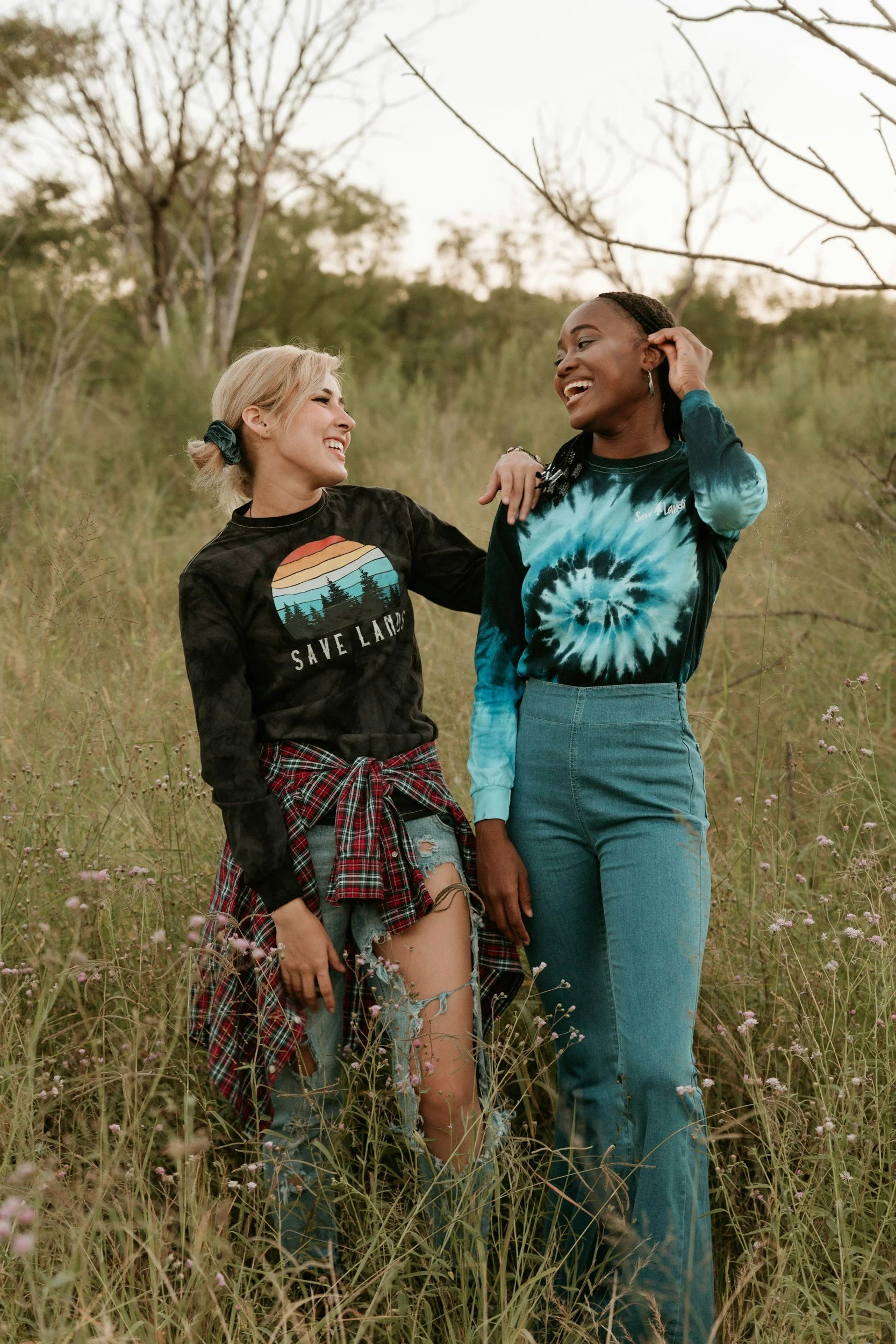 Two women laughing and smiling in a grassy field with trees in the background. One woman has blonde hair in a ponytail and is wearing a black "Save Land" T-shirt, ripped jeans, and a red plaid shirt tied around her waist. The other woman has dark hair with braids and is wearing a blue tie-dye long-sleeve top and high-waisted jeans.