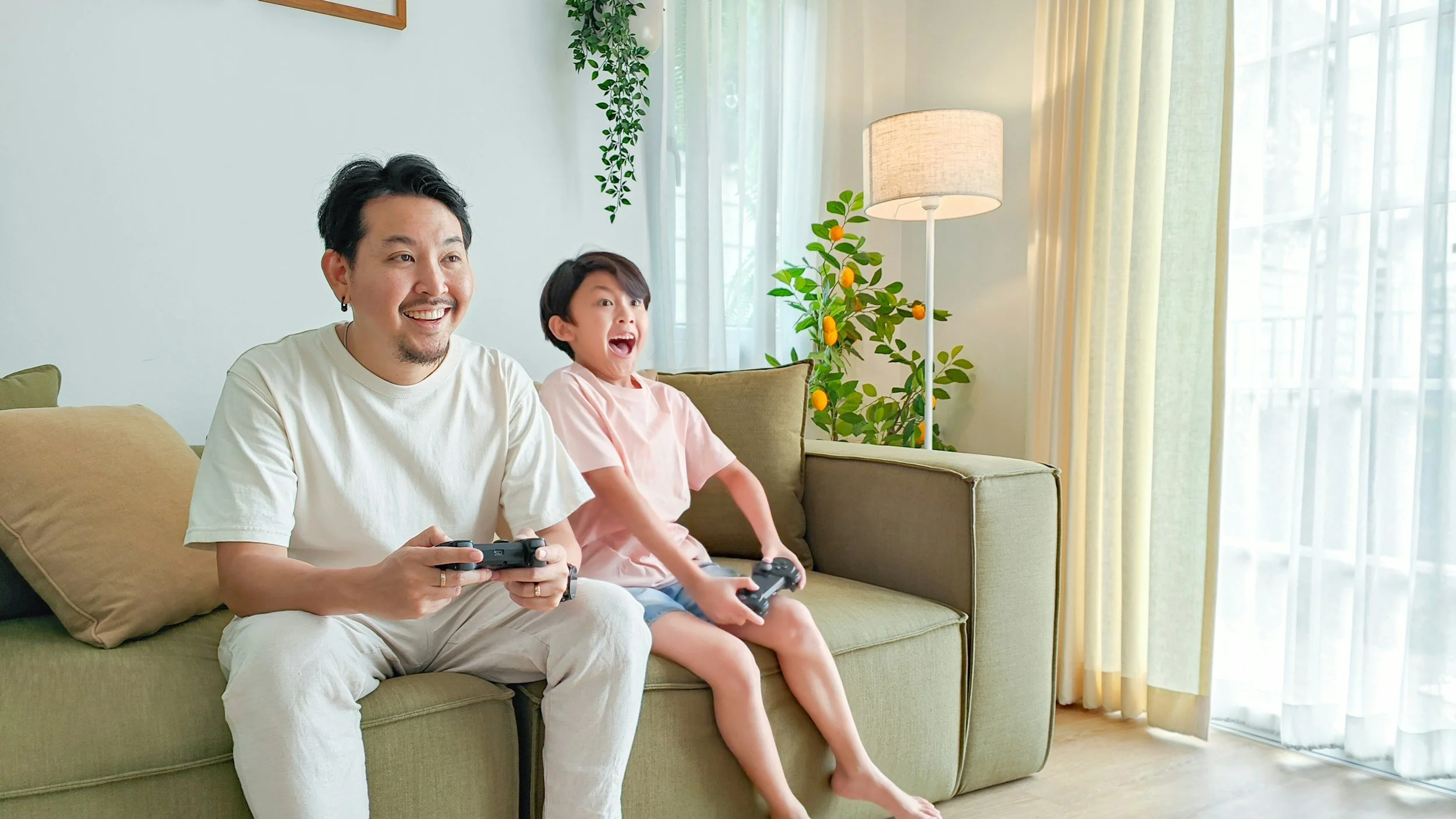 A man and a boy sitting on a green couch playing video games inside a bright living room.