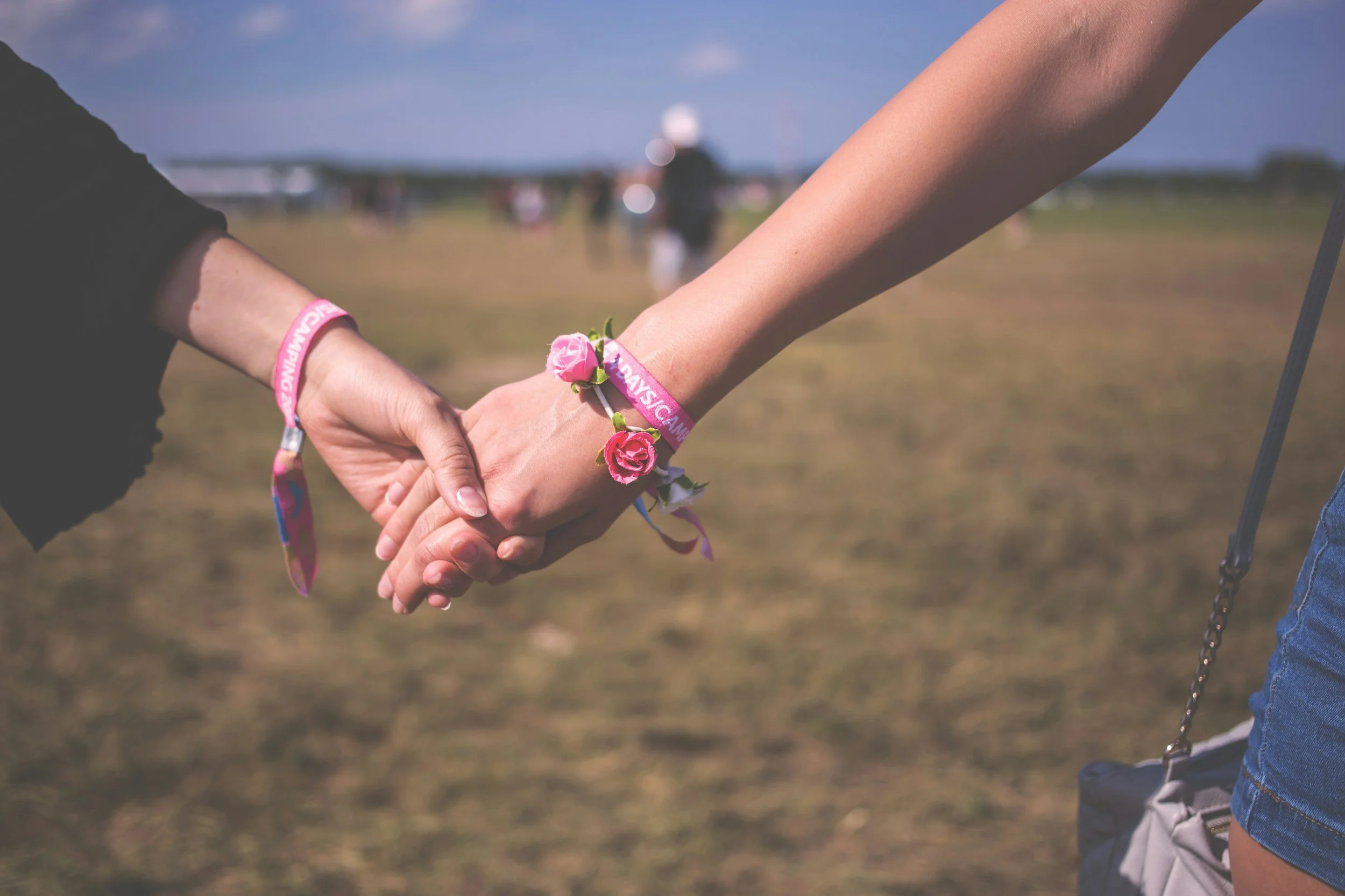 Two people holding hands at an outdoor event, both wearing pink wristbands and flower bracelets, with a blurred background of people and open sky.