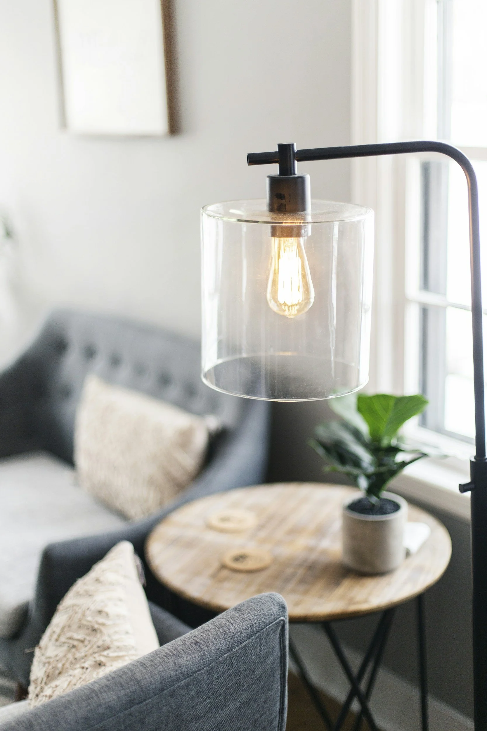 Living room with a gray couch, beige and gray pillows, a wooden sconce, and a black floor lamp with a glass lampshade containing a vintage-style Edison bulb, next to a coffee table and a potted plant near a window with blinds.