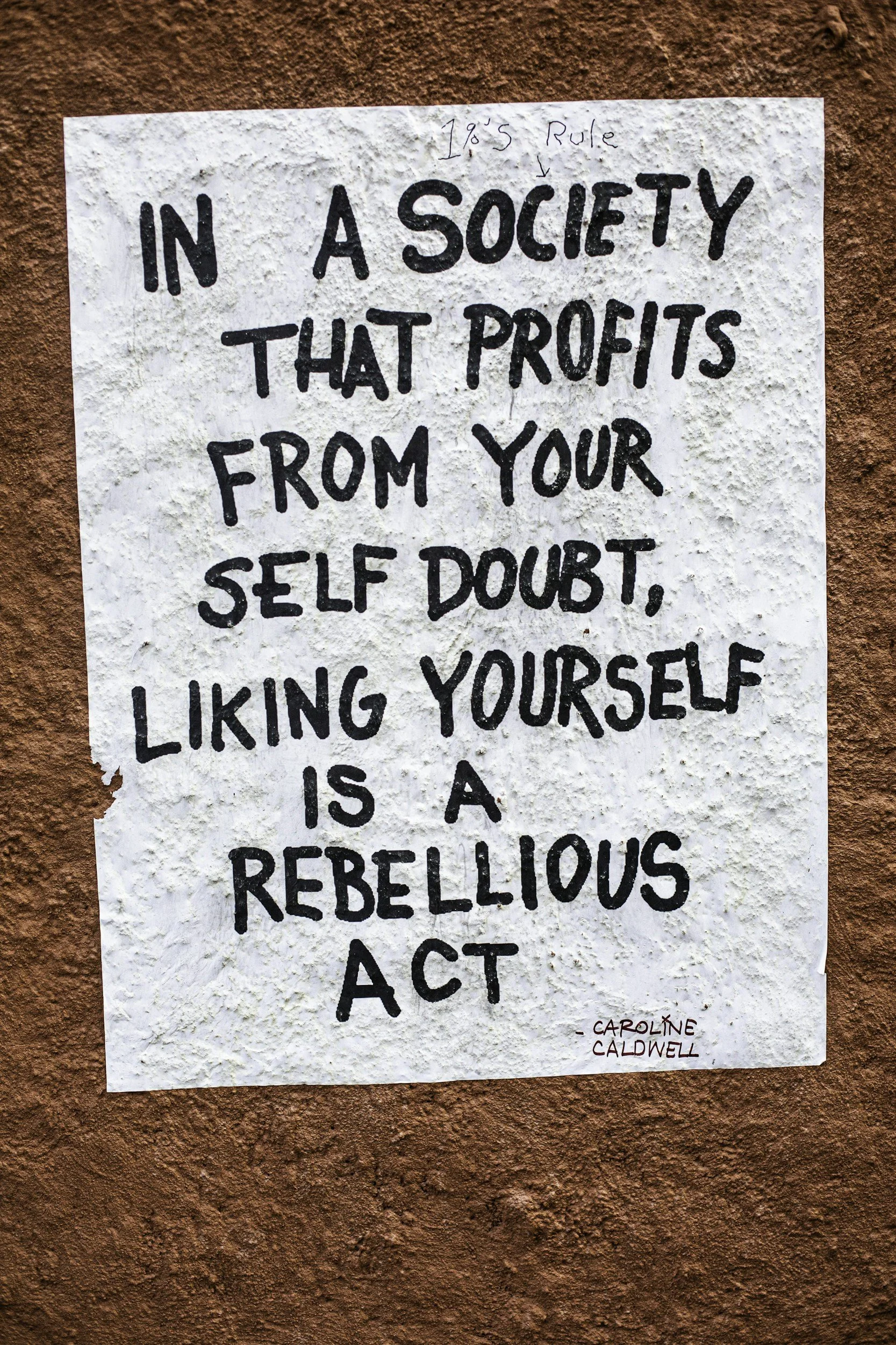 Handwritten sign on textured white paper with the quote, 'In a society that profits from your self doubt, liking yourself is a rebellious act,' attributed to Caroline Caldwell. The sign is taped to a reddish-brown wall.