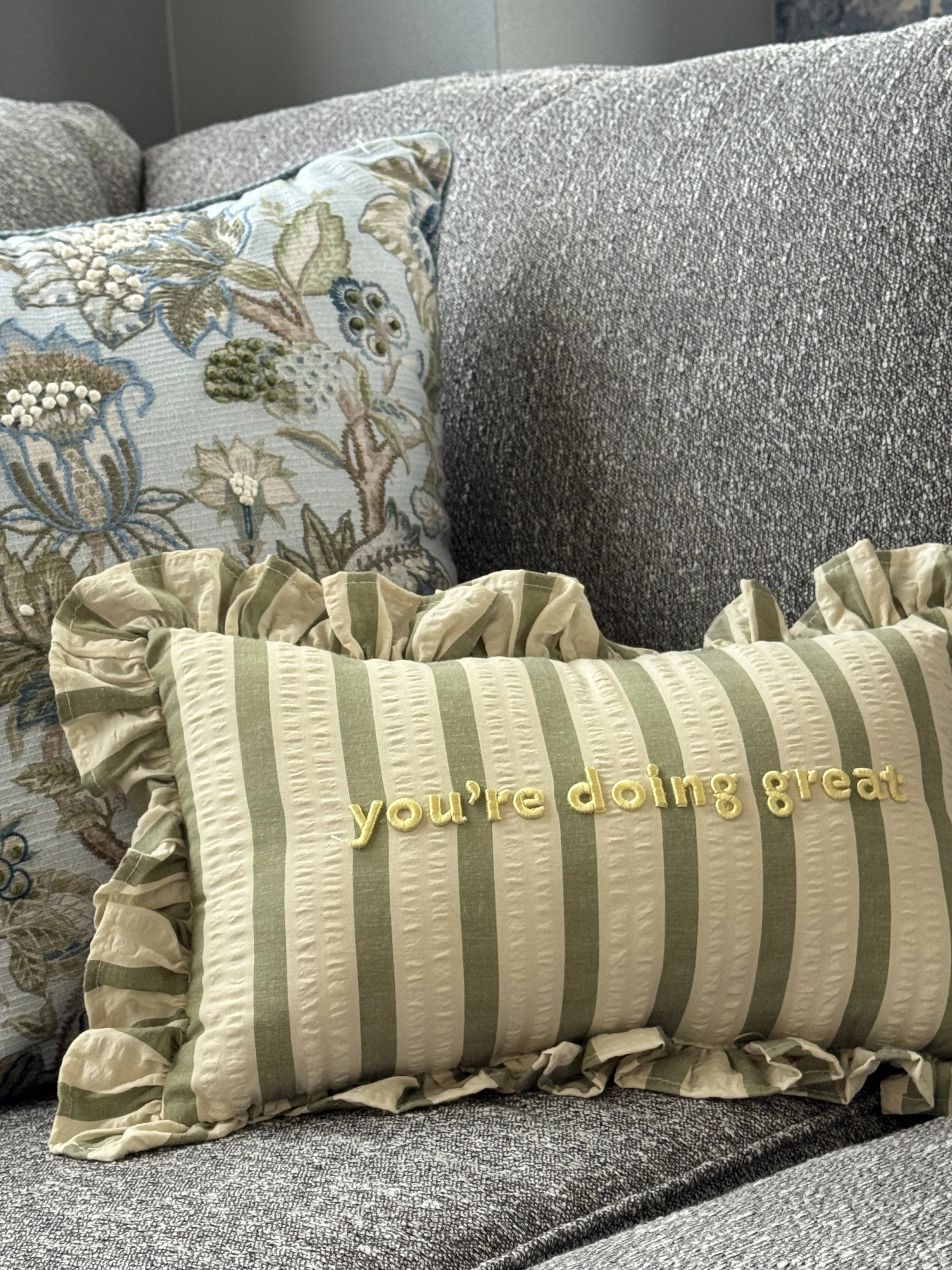 Decorative pillows on a gray textured sofa, including one with yellow embroidery saying 'you're doing great' and ruffled edges, and another with floral embroidery and a ruffled edge.