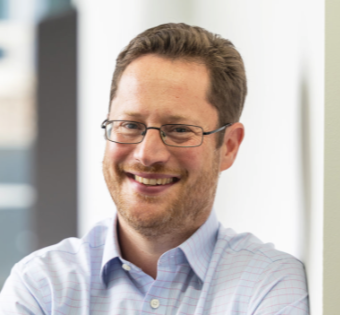 Man with glasses smiling while leaning against a wall in a professional setting.