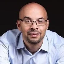 A person wearing glasses and a light blue shirt, leaning forward with arms crossed on a chair, posing against a black background.