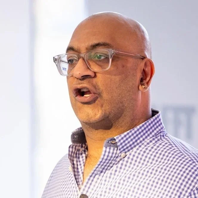 Man speaking in a blue checkered shirt, wearing glasses and a microphone on his collar.