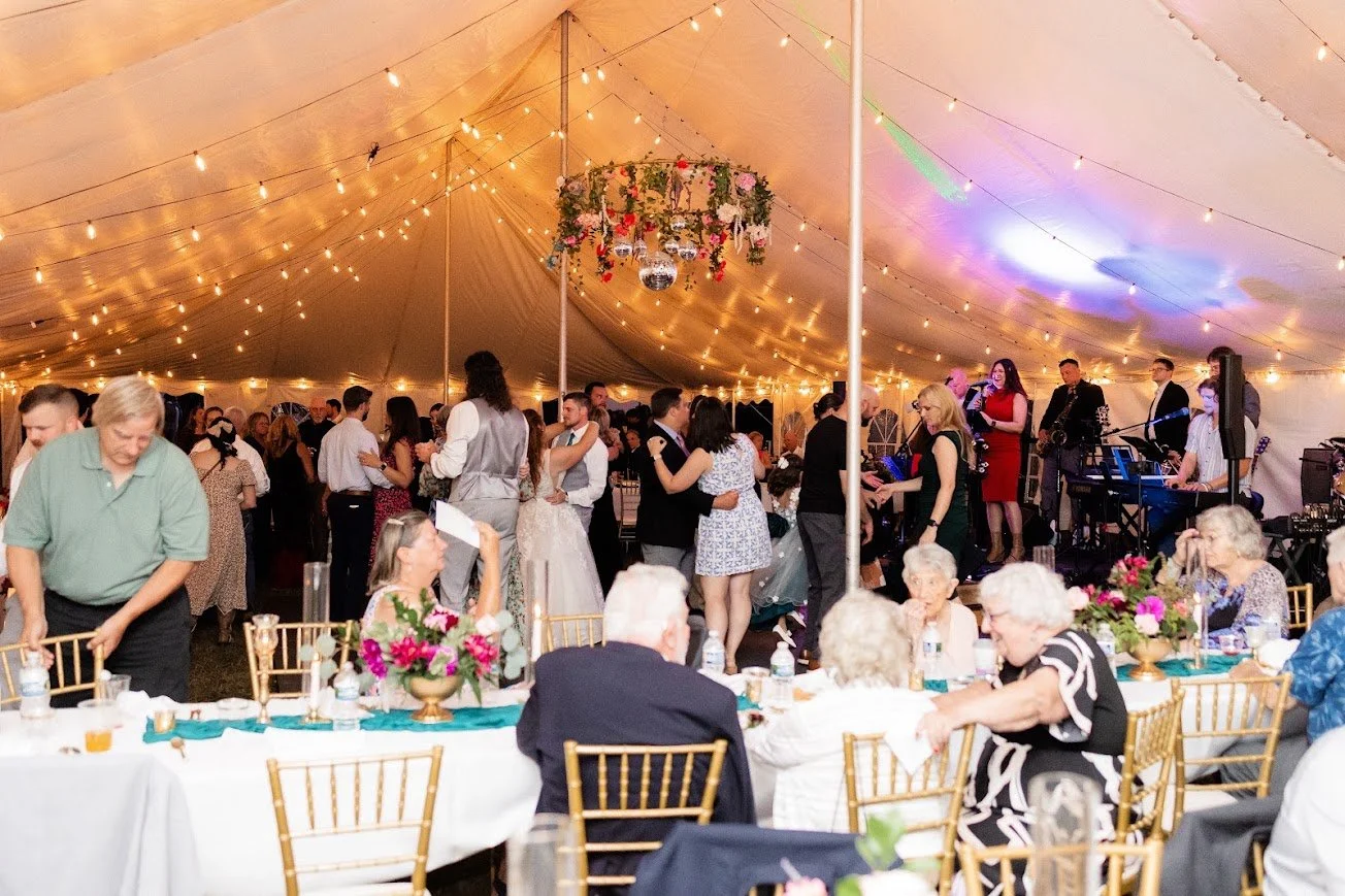 Outdoor evening setting with five high tables draped in white cloths, decorated with small floral arrangements, bottles, and glasses, on a grassy lawn under string lights between tall trees.
