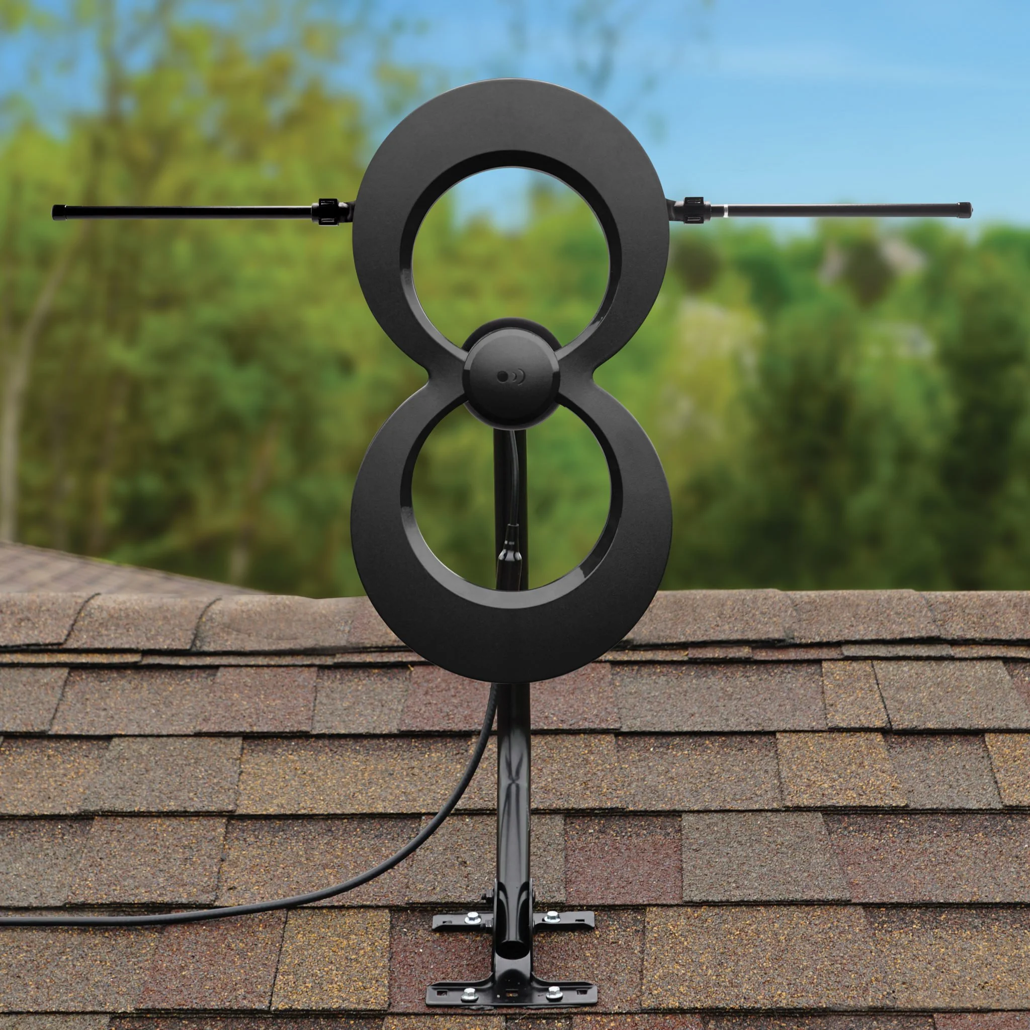 A rooftop-mounted TV antenna with a circular design and horizontal rods, installed on a shingled roof with a blurred green forest and blue sky background.