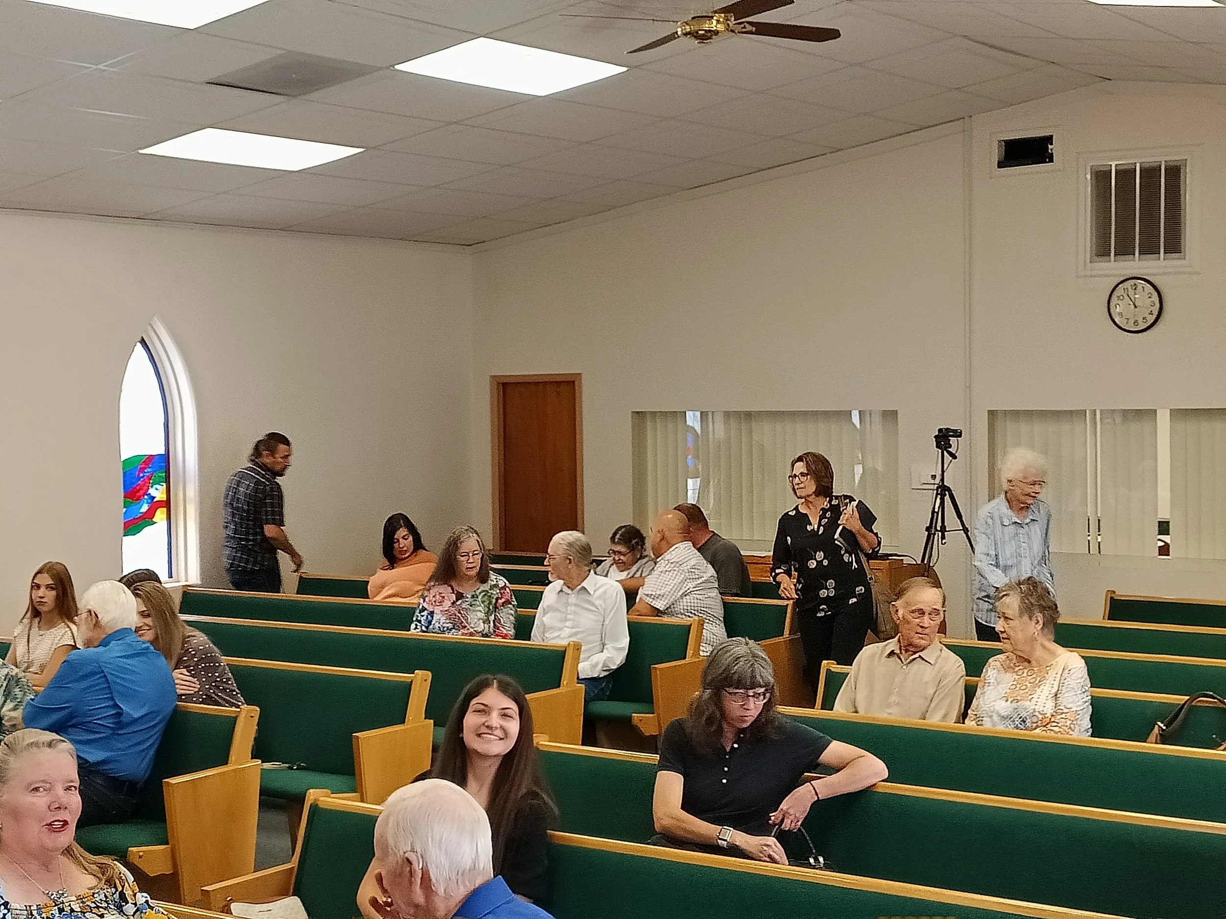 A group of people gathered in a church or community hall, seated on green pews. Some are engaging in conversations, while others are listening. The room has a high ceiling with ceiling fans, a clock showing 4:09, and a stained-glass window on the left.