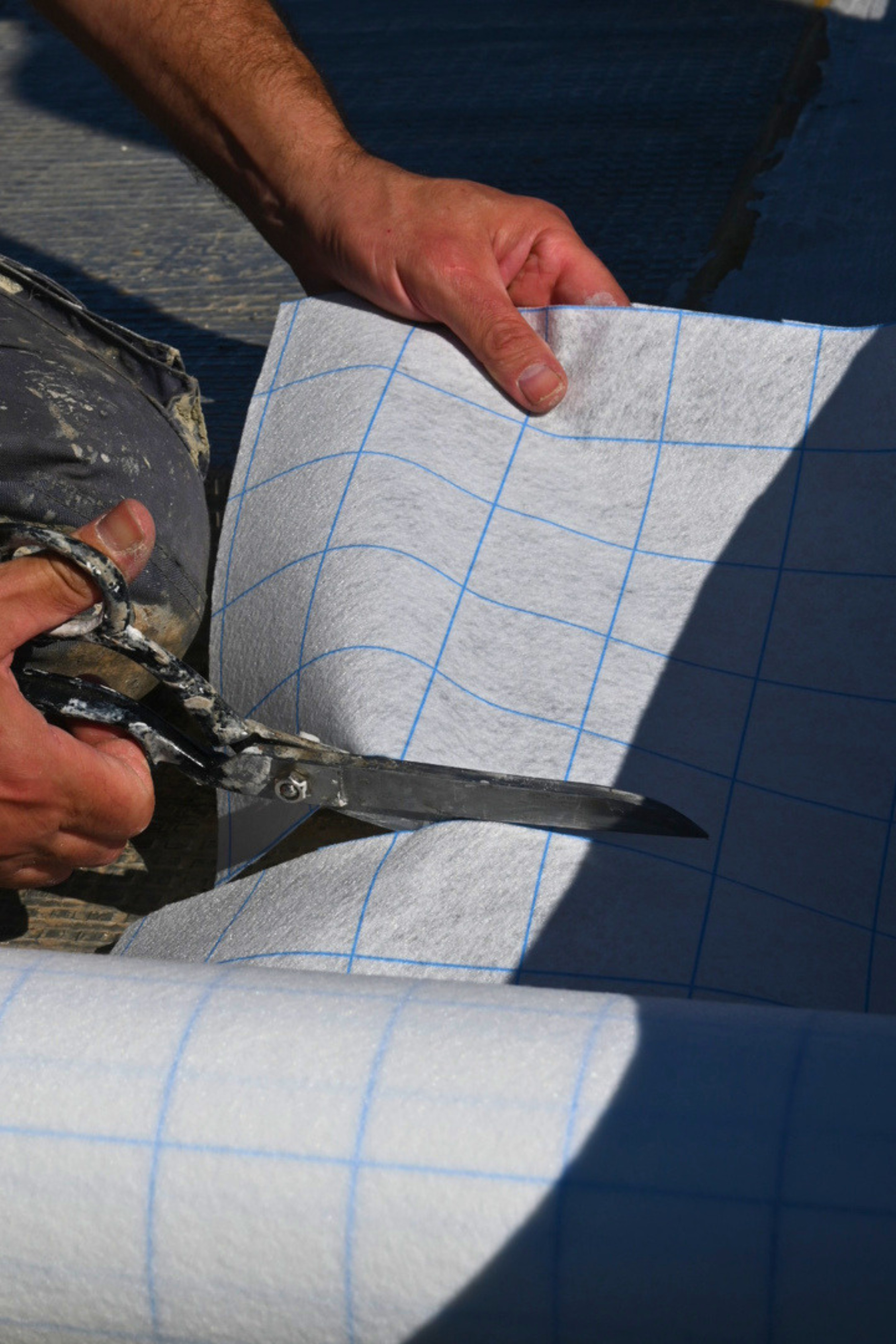 Une personne coupe une feuille de matériau isolant blanc avec un motif de lignes bleues, utilisant une paire de ciseaux, sous la lumière du soleil.