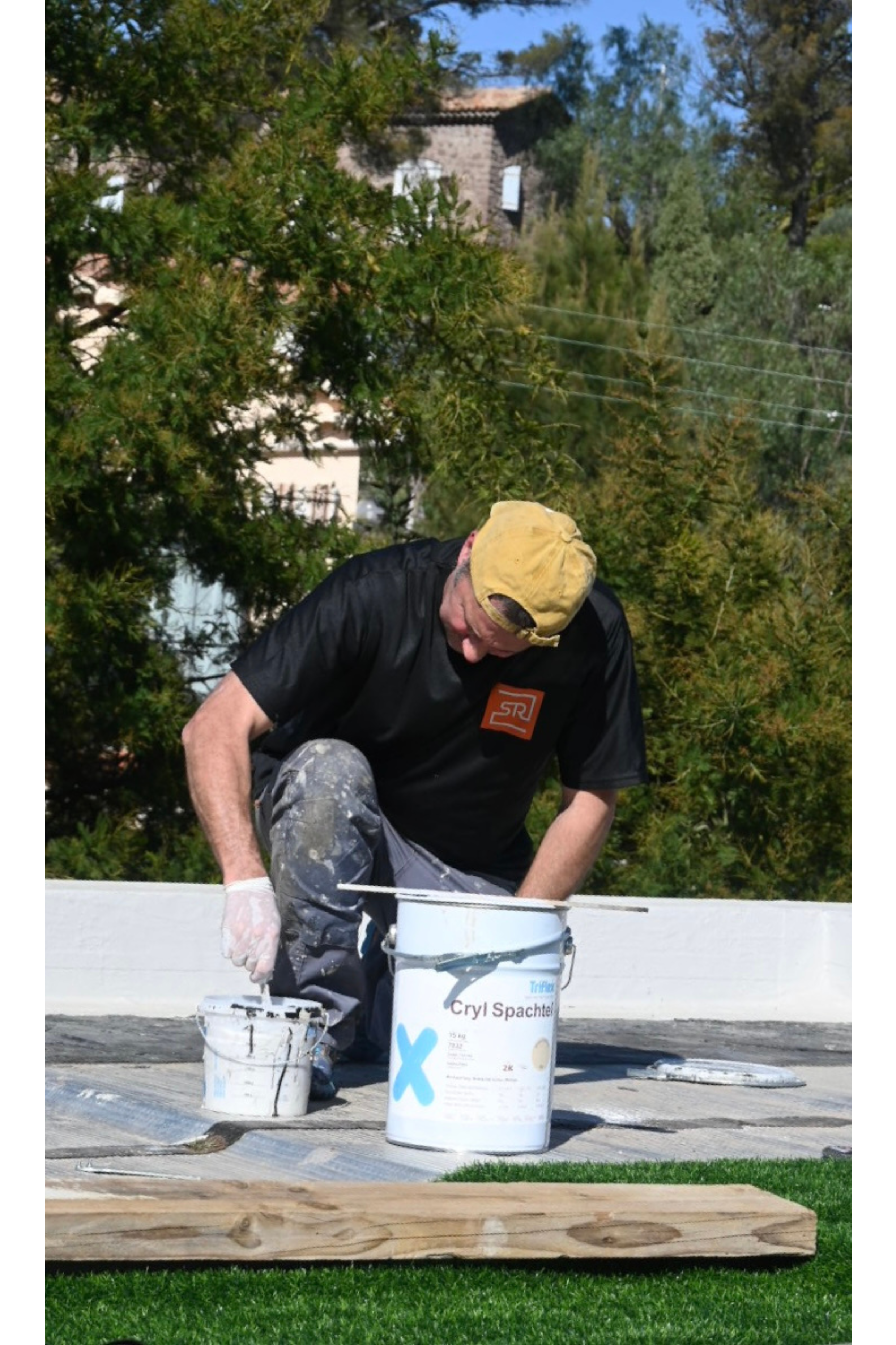 Un homme en train de peindre une toiture avec de la résine d'étanchéité liquide blanche, portant un chapeau jaune et des vêtements de travail sales, dans un environnement extérieur avec des arbres en arrière-plan.
