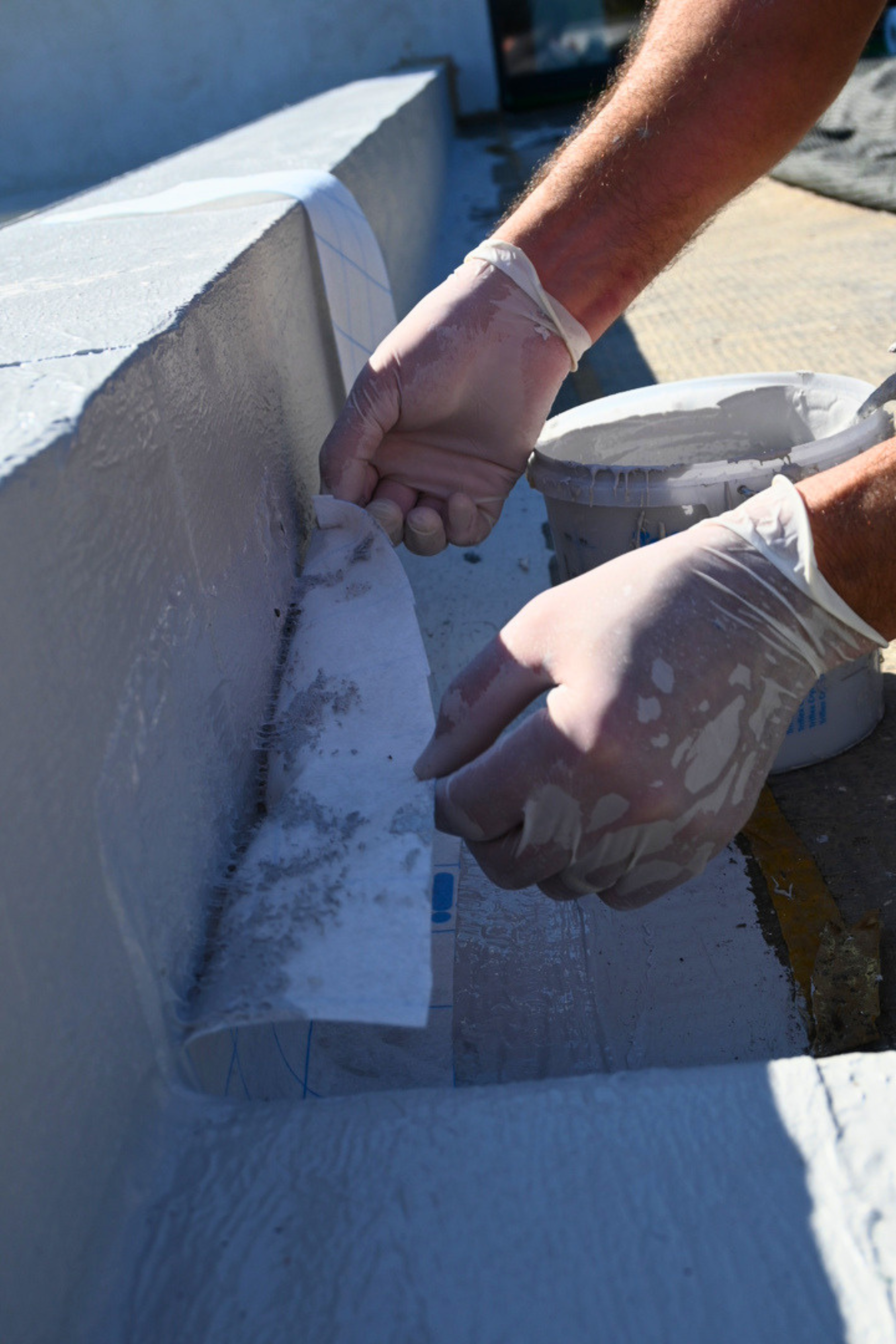 Une personne en gants applique de la résine d'étanchéité liquide blanche sur un mur en utilisant une spatule.