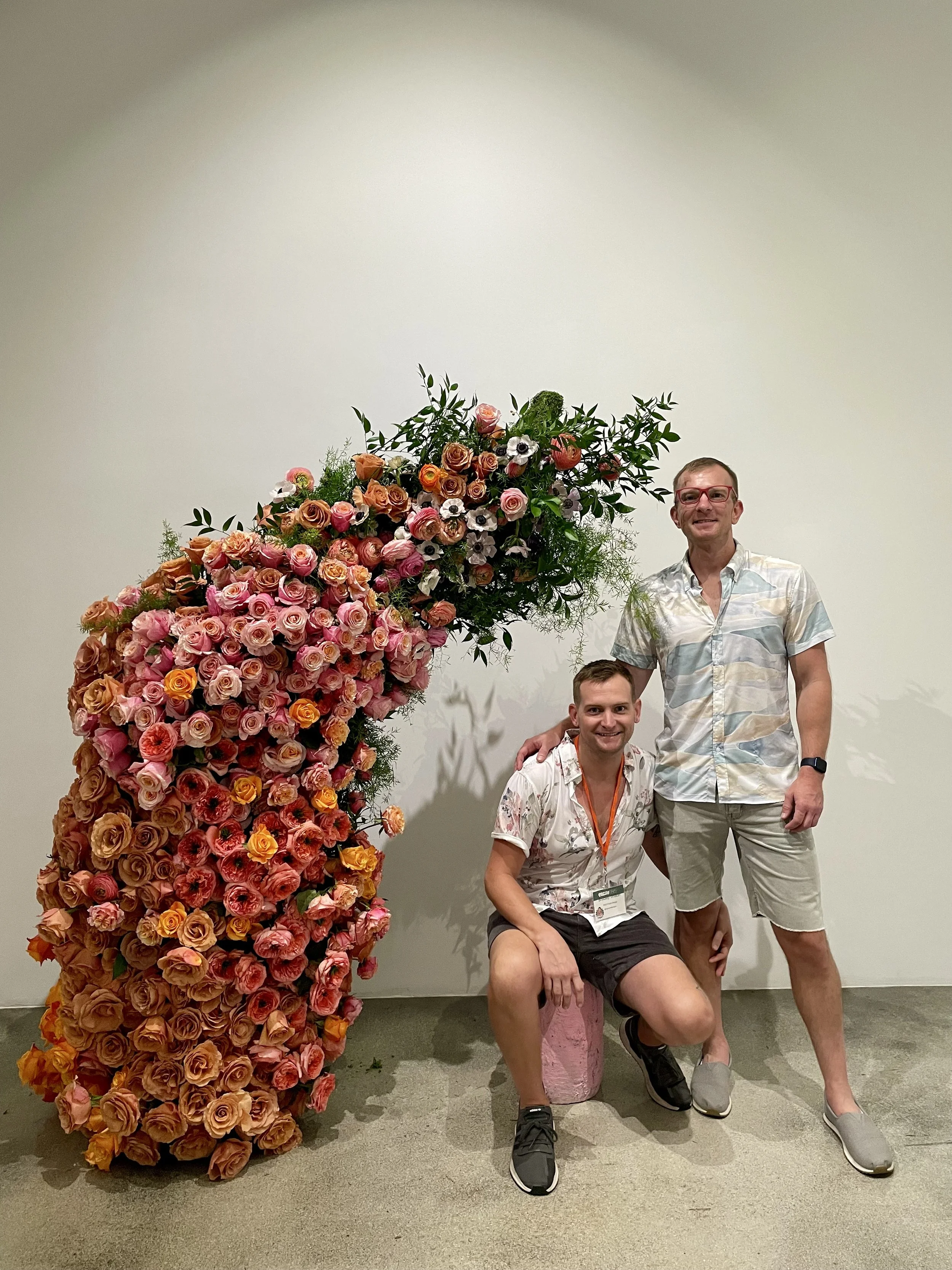 Two smiling men with casual summer clothing and glasses pose next to a large flower arrangement of pink, orange, and white roses with green foliage in a gallery or event space with plain white wall.