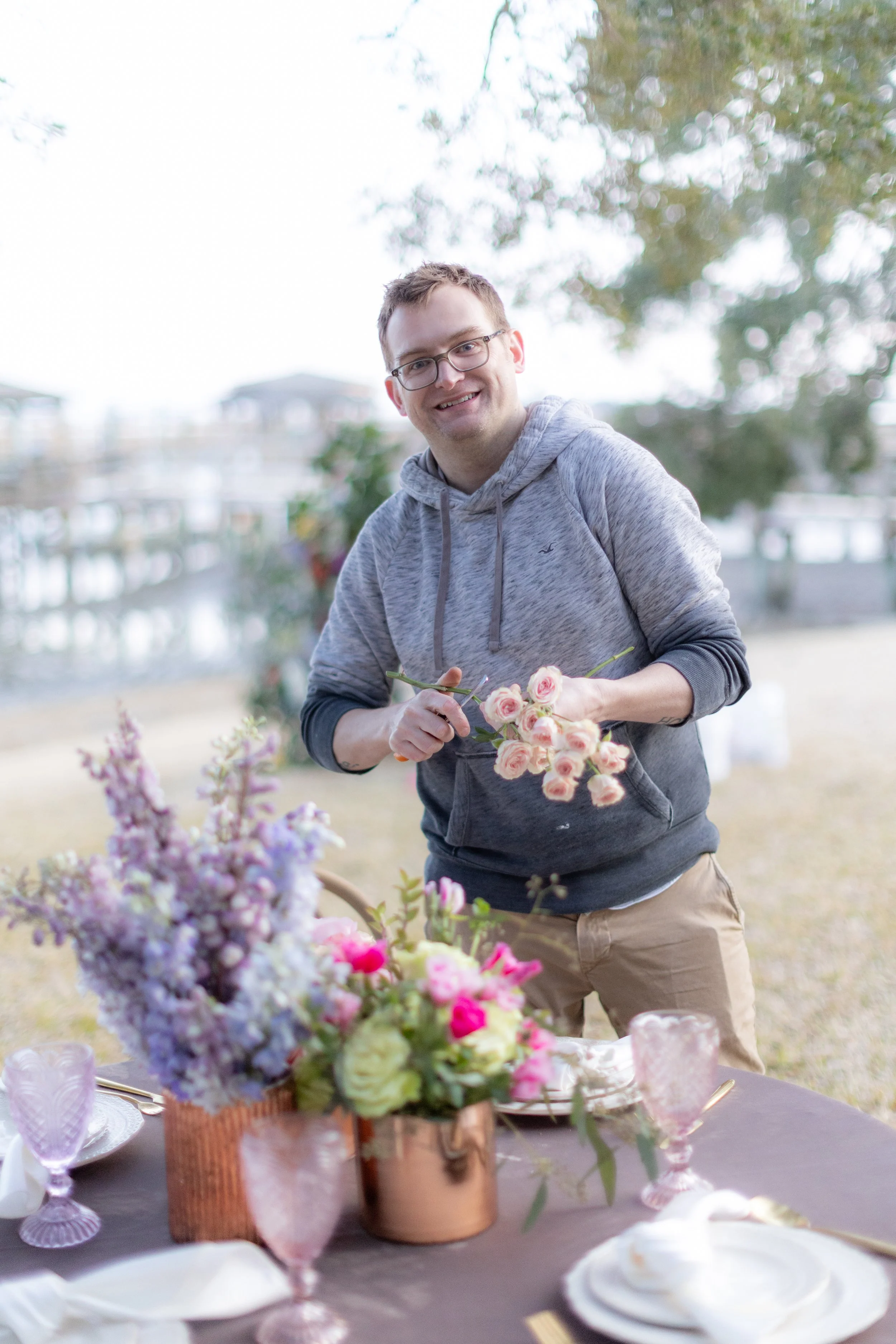 A young man with glasses and a gray hoodie is smiling and holding a bouquet of pale pink roses. He is outdoors, near a table with a floral centerpiece, plates, and glasses, with a blurred background of trees and a walkway.