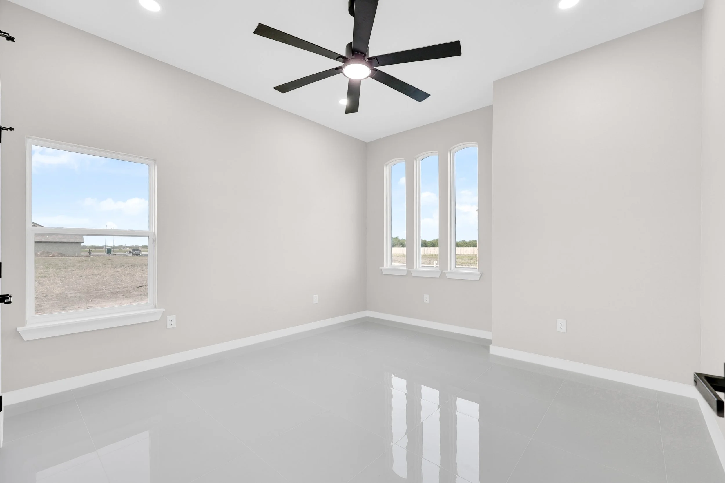 Empty room with light gray walls, glossy tile floor, large black ceiling fan, and two windows with natural light.
