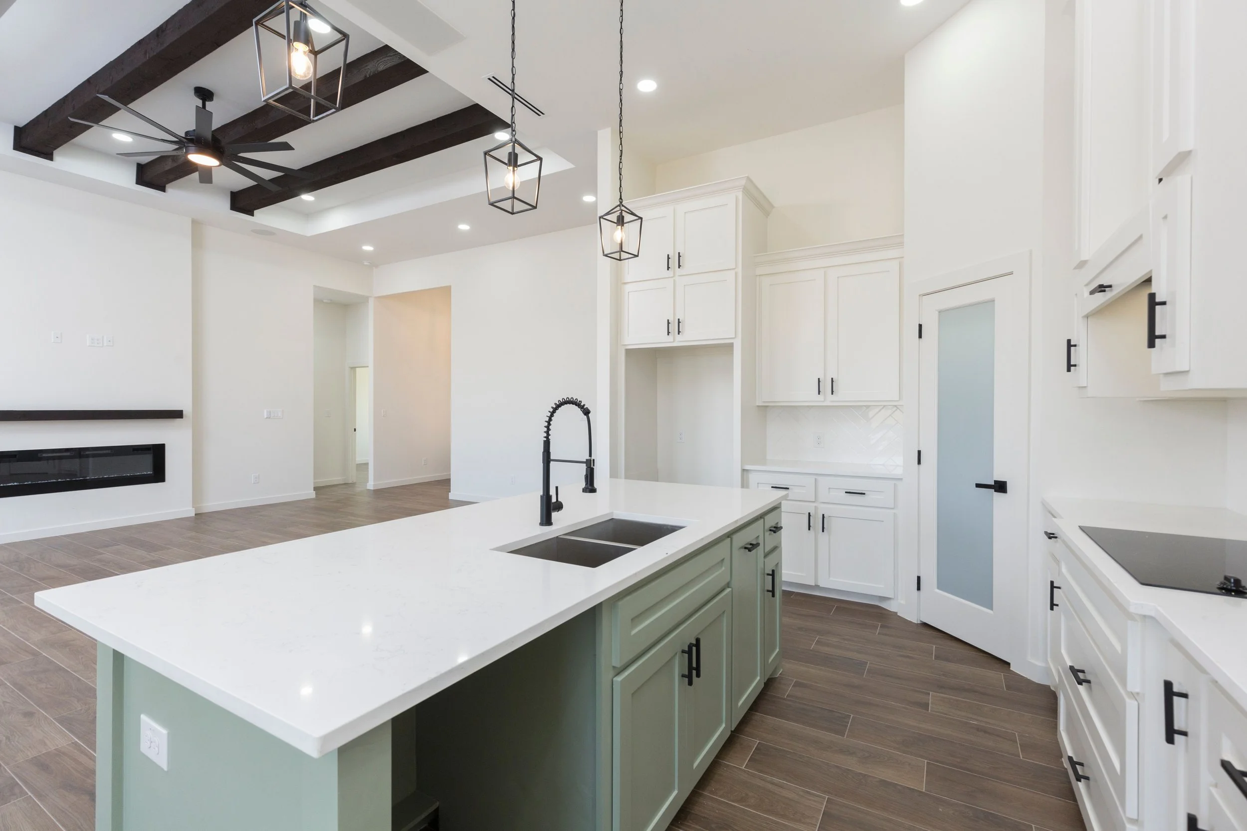 Modern kitchen interior with white cabinets, green island, black fixtures, wooden flooring, and ceiling beams. Features pendant lighting and built-in stove.
