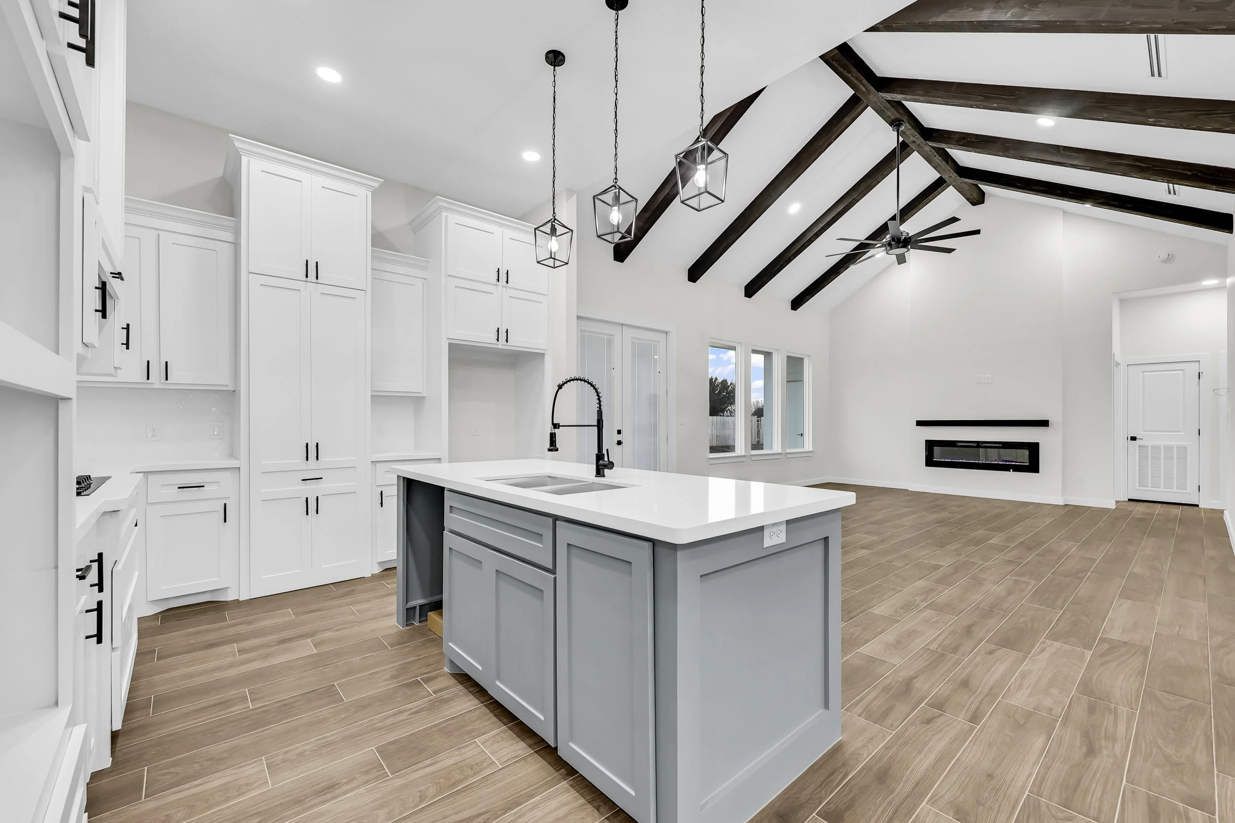 Modern kitchen and living area with white cabinets, wood beam ceiling, island with sink, pendant lights, and hardwood flooring.