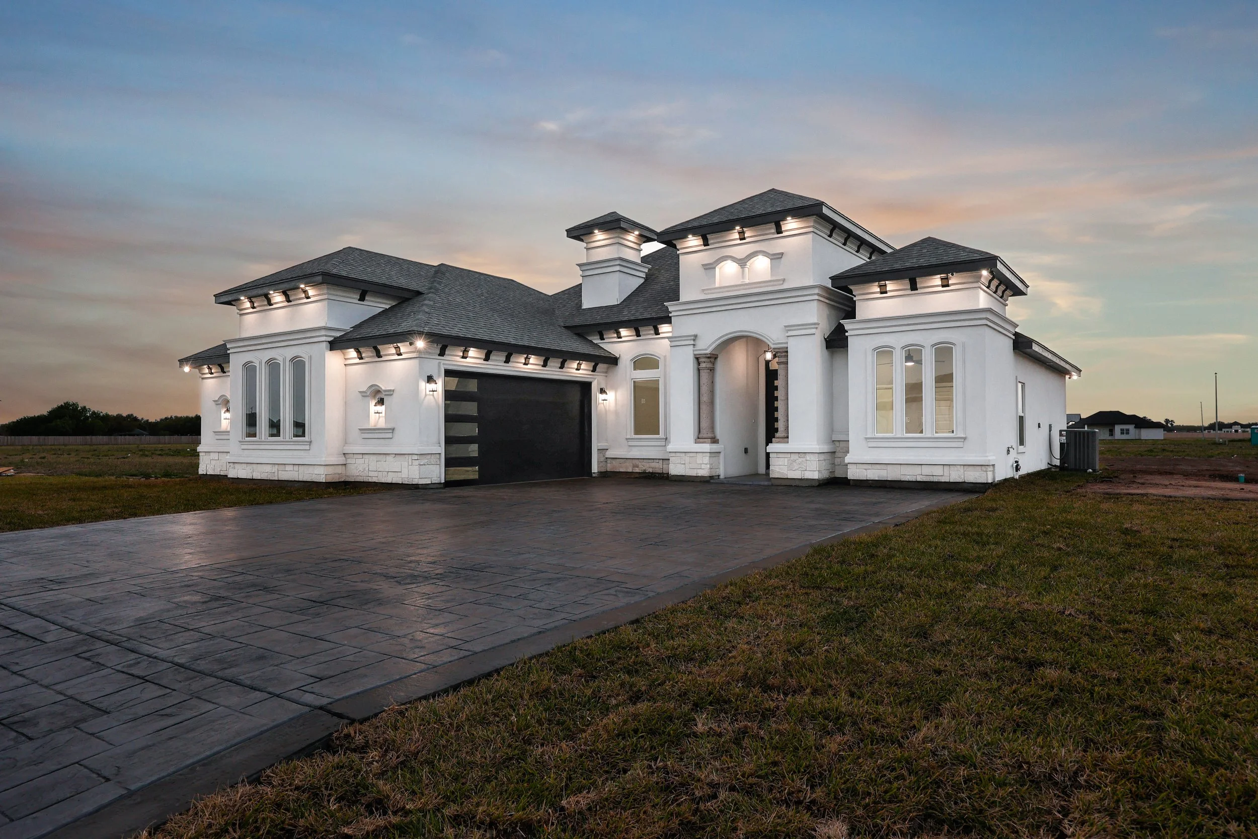 Modern white house with gray roof, large driveway, and decorative exterior lighting at dusk.