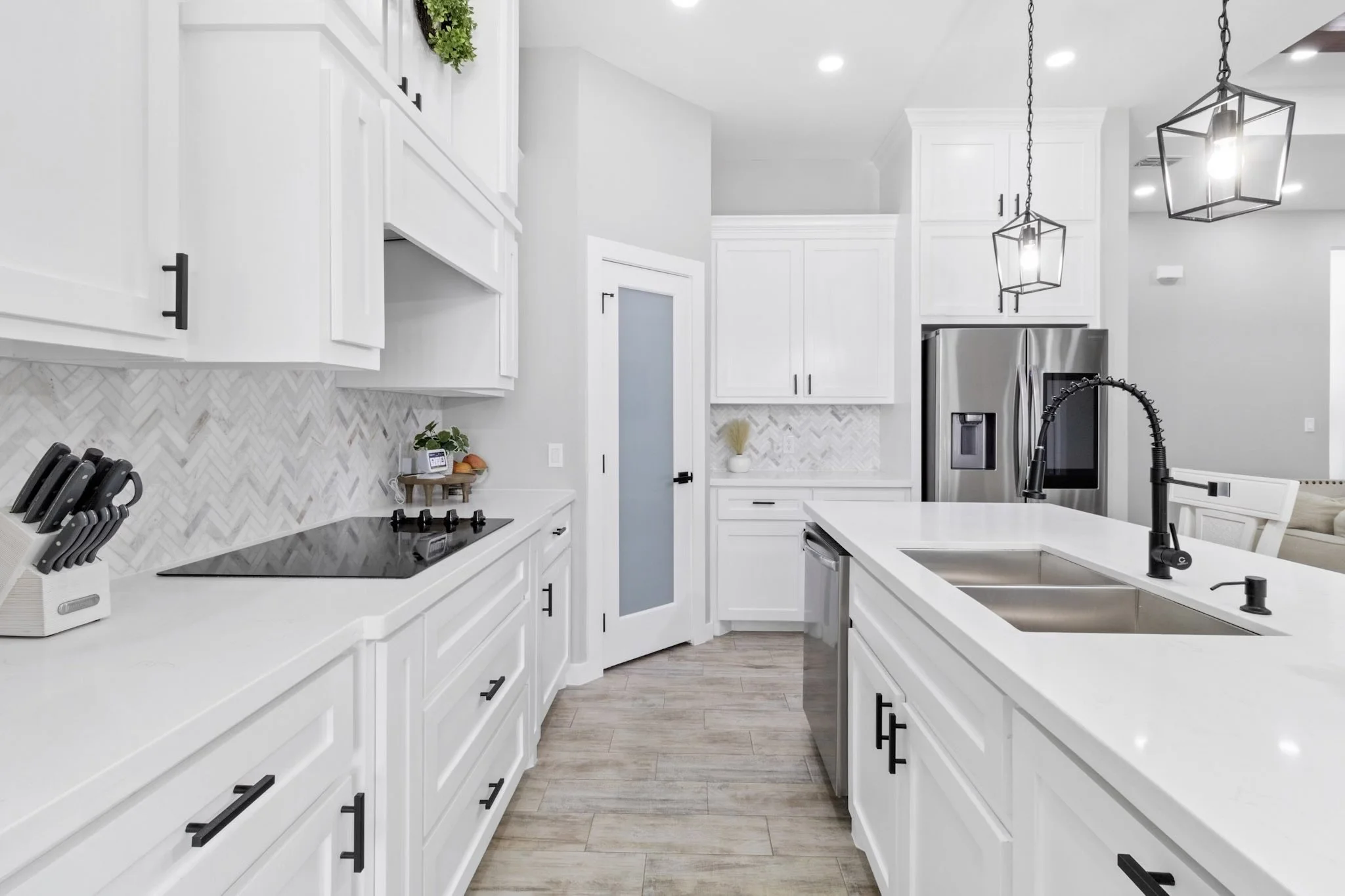 Modern white kitchen with marble backsplash, black countertop stove, stainless steel appliances, double sink, and pendant lights.