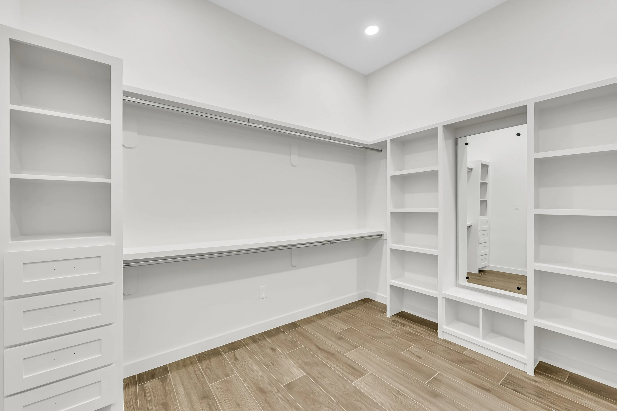 Spacious empty walk-in closet with white shelving units, hanging rods, and a full-length mirror, featuring wood-look tile flooring.