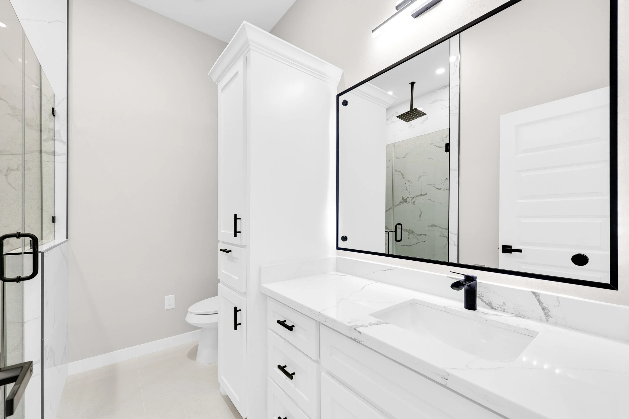 Modern white bathroom with a large mirror, black fixtures, and a marble countertop. Features a shower with glass doors, a toilet, and a cabinet.