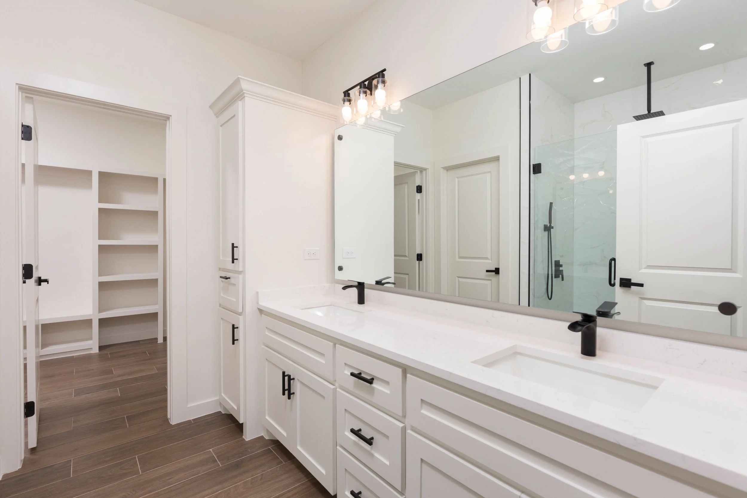 Modern bathroom with double vanity, black fixtures, large mirror, light fixtures, white cabinets, wood flooring, and adjoining closet and shower area.
