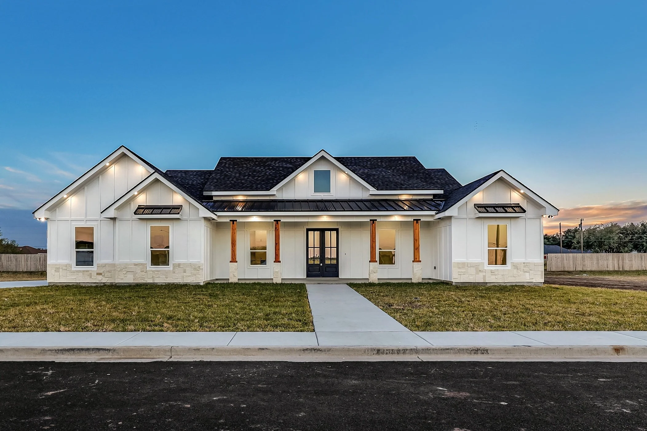 Modern white farmhouse-style house with a black roof, featuring multiple gables and a central front entrance, viewed during dusk.