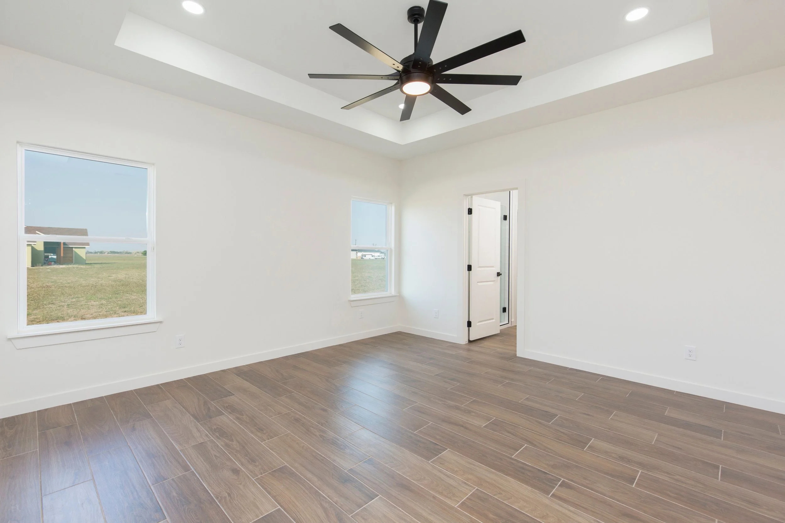 Empty room with wood flooring, ceiling fan, two windows, and an open door to another room.