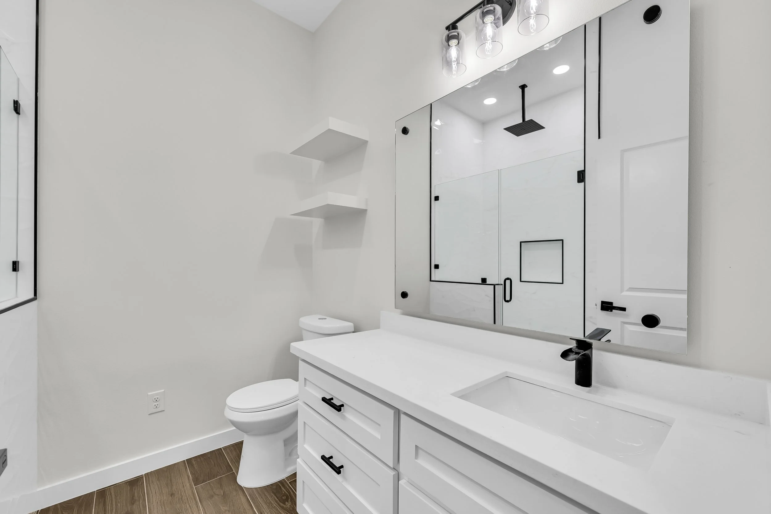 Modern white bathroom with a large mirror, a black faucet on a white countertop, white cabinets, a toilet, wall-mounted shelves, and a glass shower enclosure with black trim.