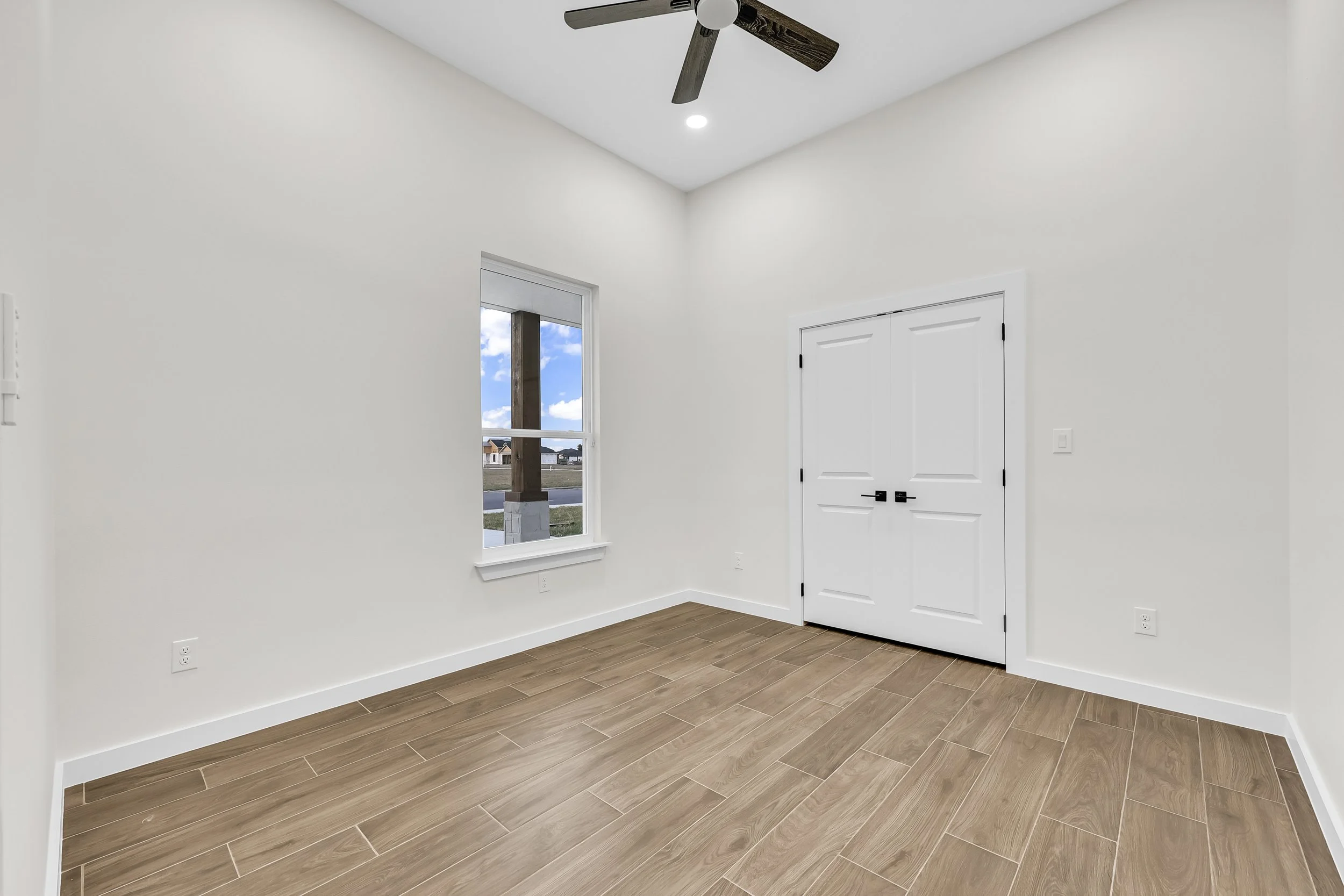 Empty room with wooden tile flooring, white walls, a window, a ceiling fan, and double doors.