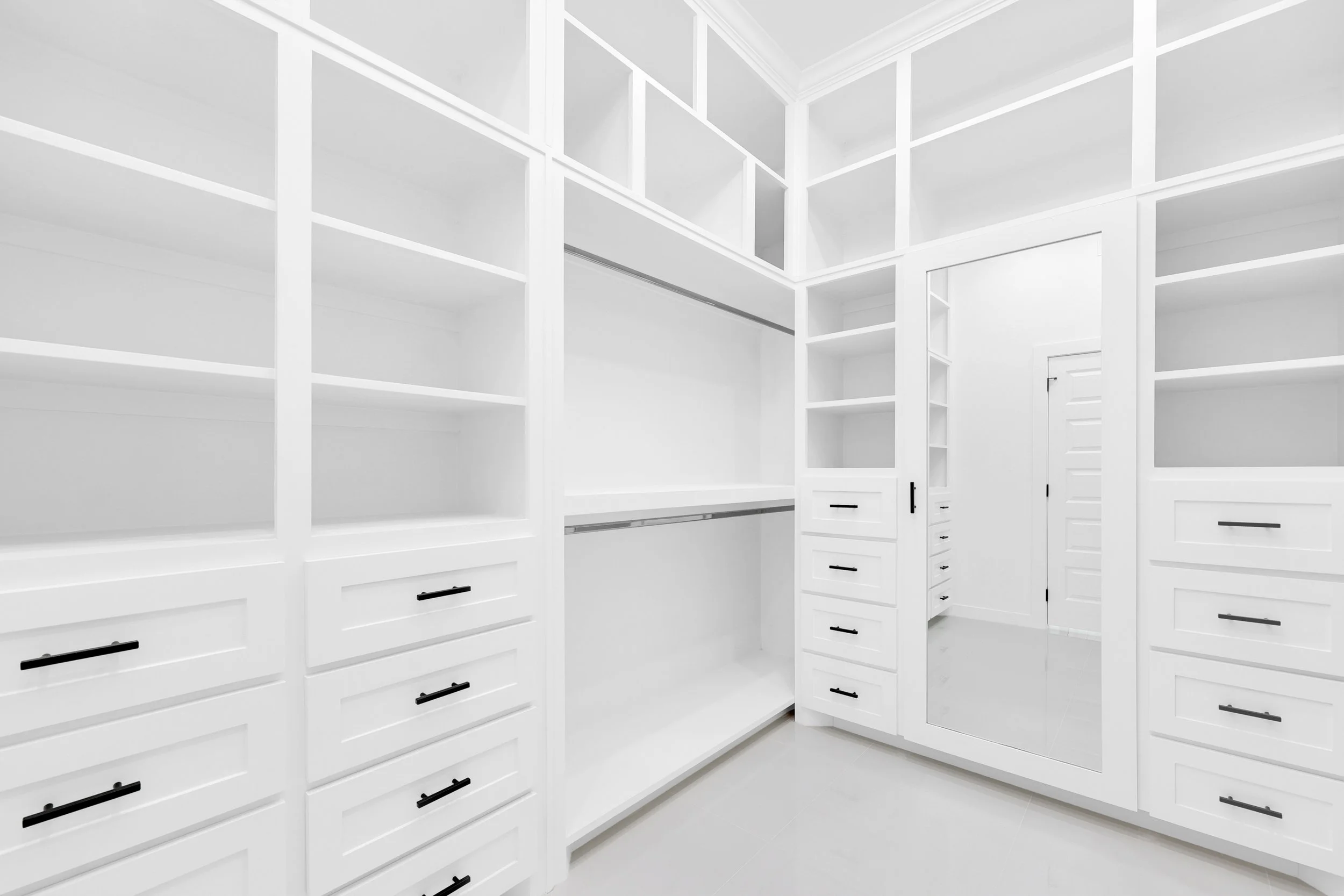Spacious modern white closet with open shelving, built-in drawers with black handles, and a large mirror.