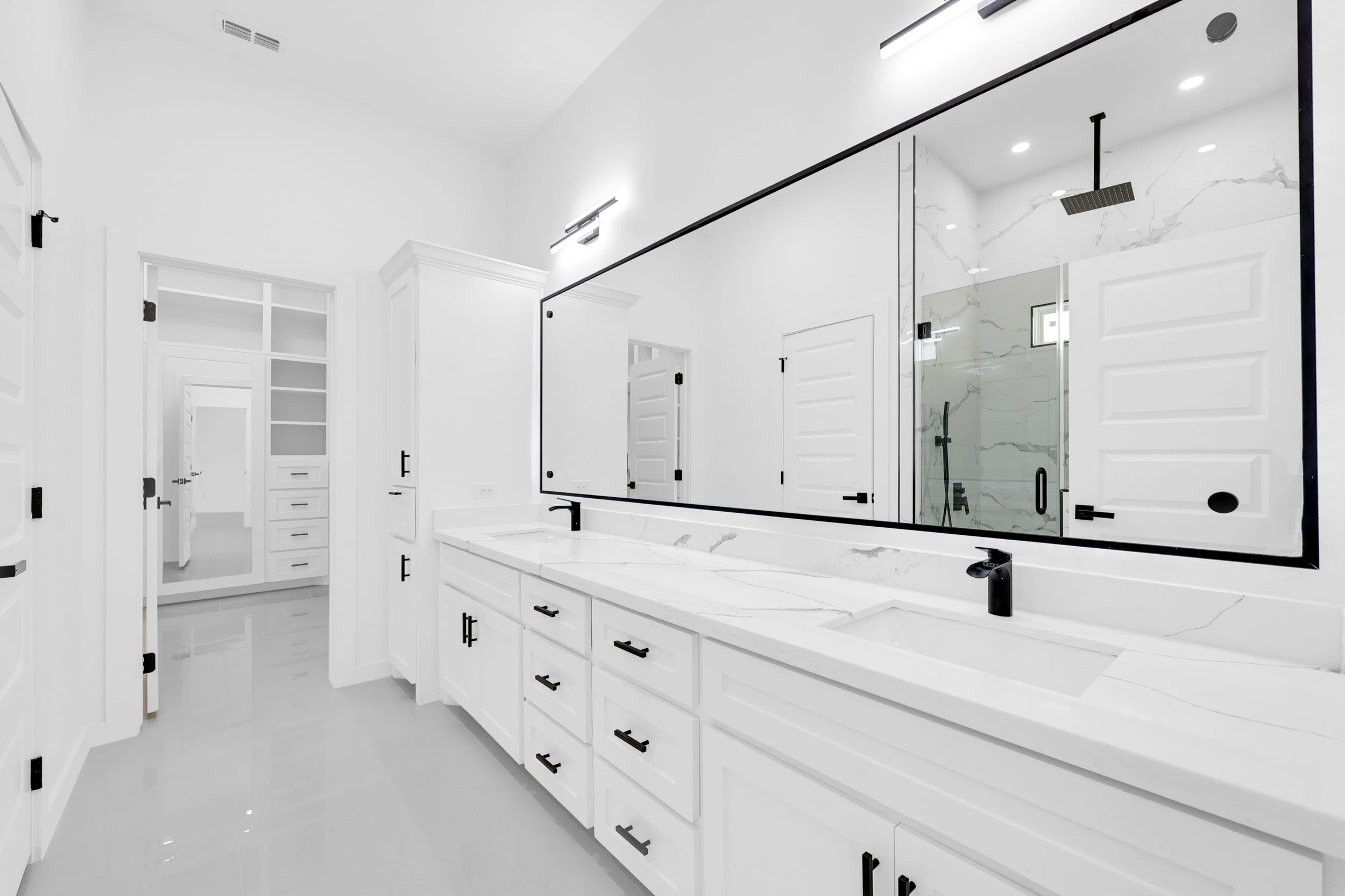 Modern white bathroom with dual sinks, large mirror, and a glass-enclosed shower with black fixtures.