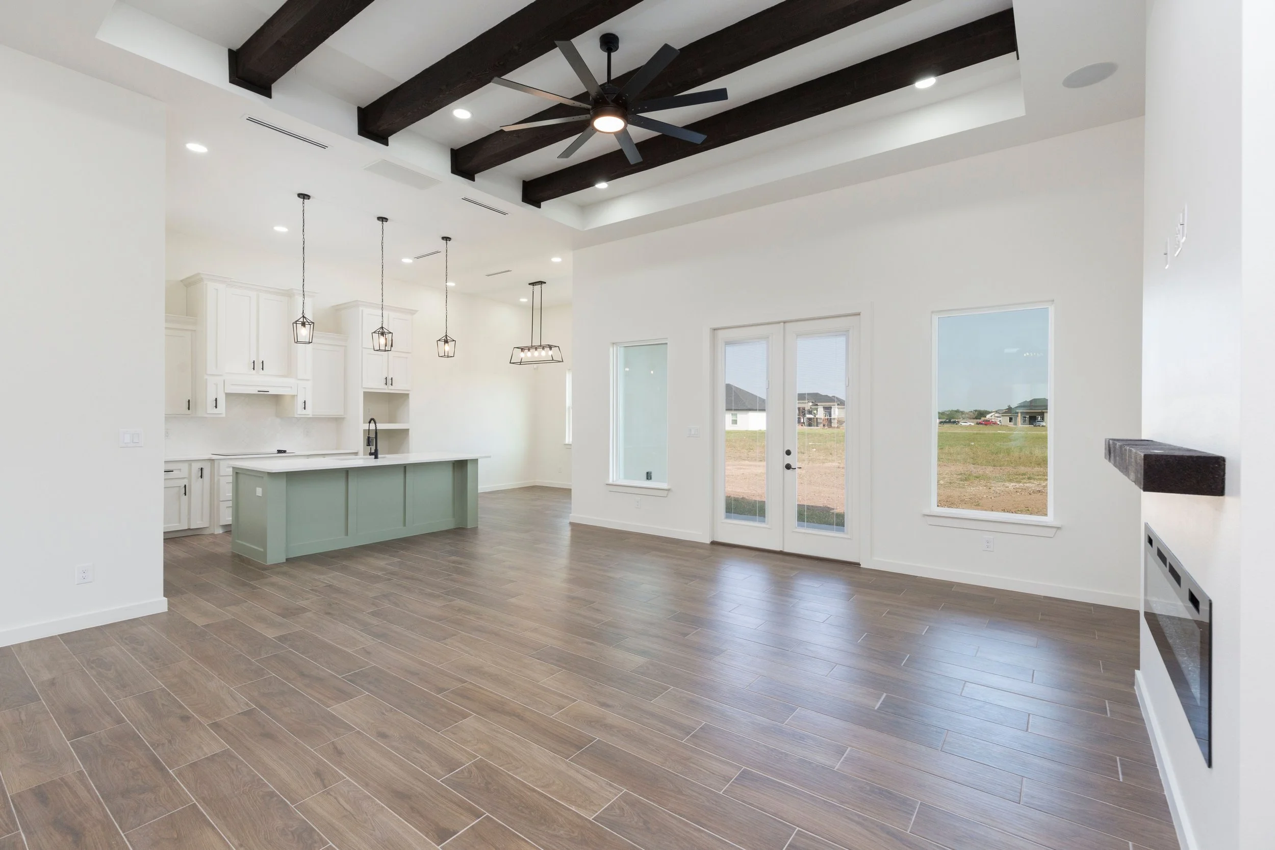 Modern open-concept living room with hardwood flooring, a kitchen island, white cabinets, pendant lights, exposed ceiling beams, large windows, and a glass door leading outside.