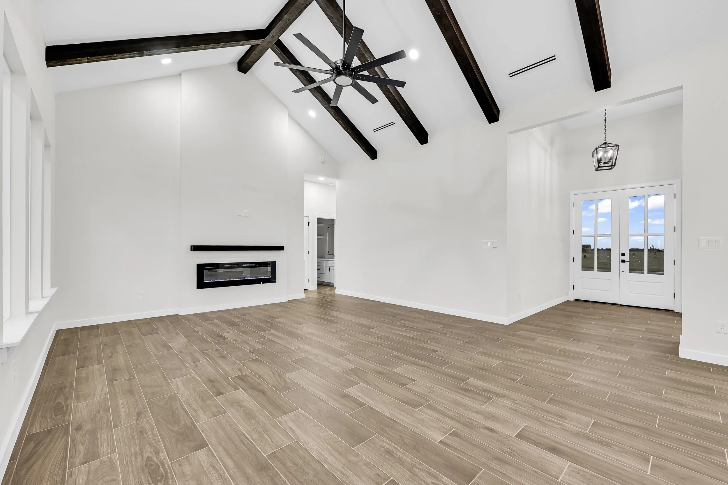 Modern interior with wood tile flooring, white walls, black ceiling beams, large ceiling fan, wall-mounted fireplace, and double doors with glass panels.