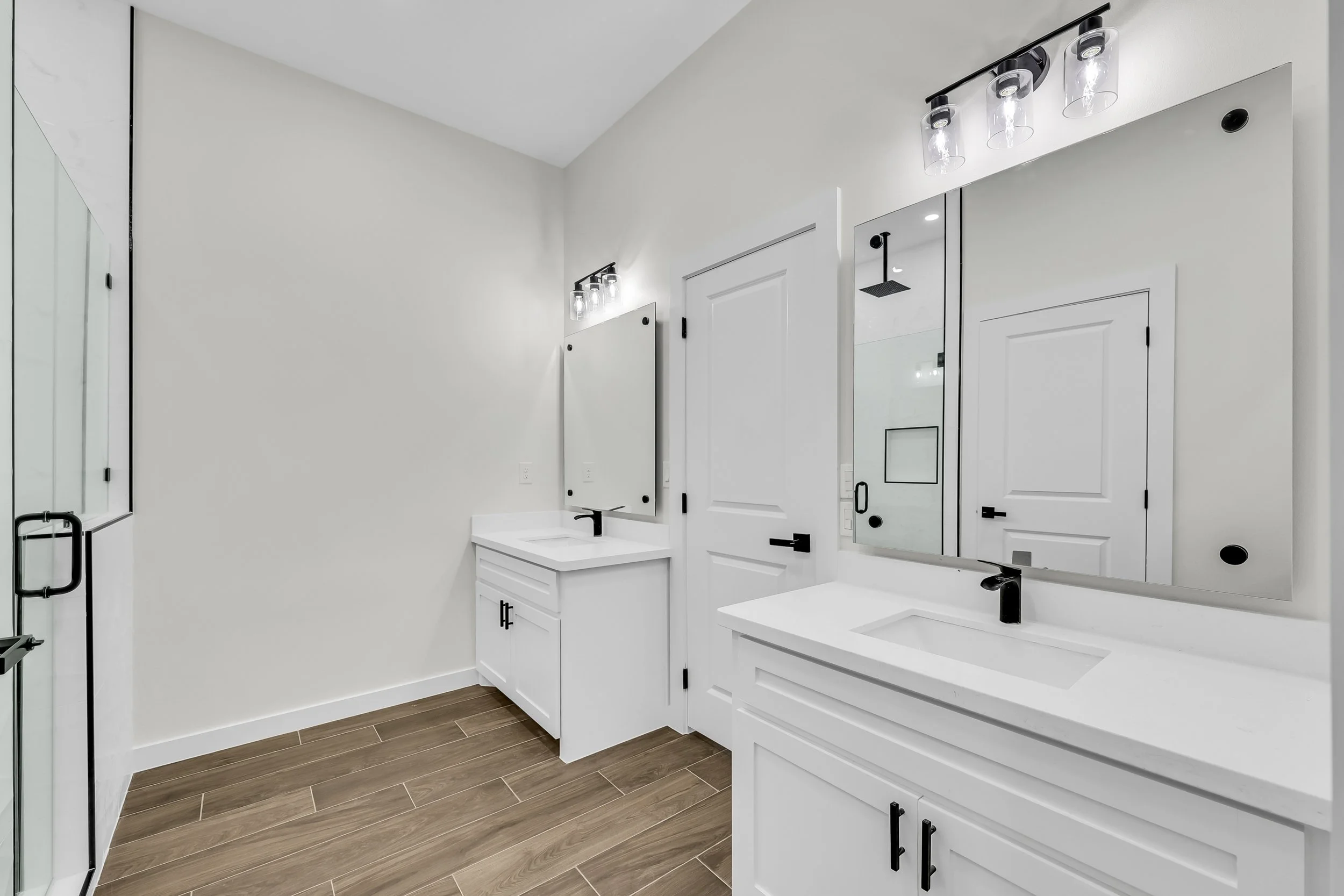 Modern bathroom interior featuring double sinks, white cabinets, large mirrors, chrome faucets, and wood-look tile flooring. Glass shower door visible on the left.