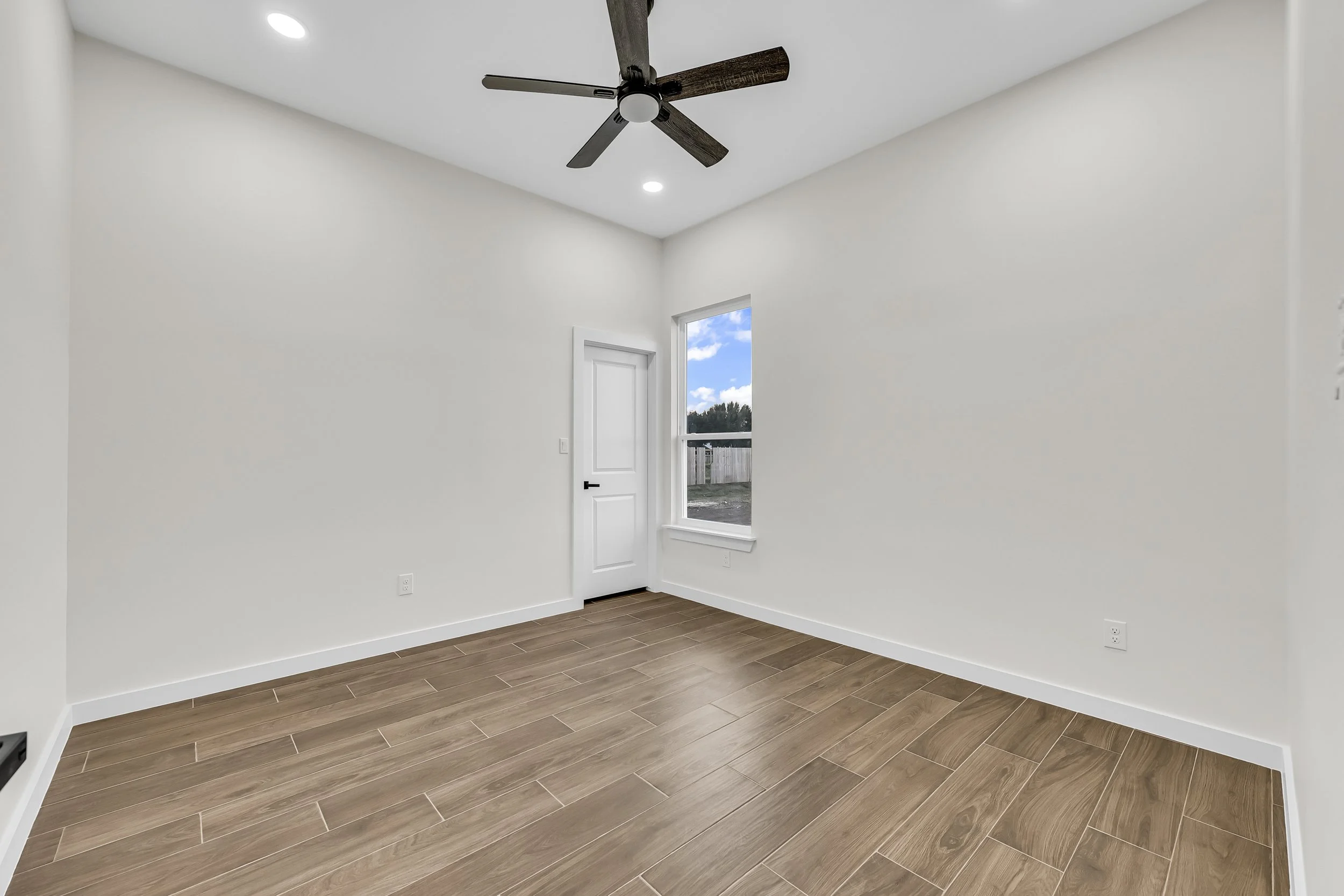 Empty room with wooden tile flooring, white walls, ceiling fan, and a window with a view outside.
