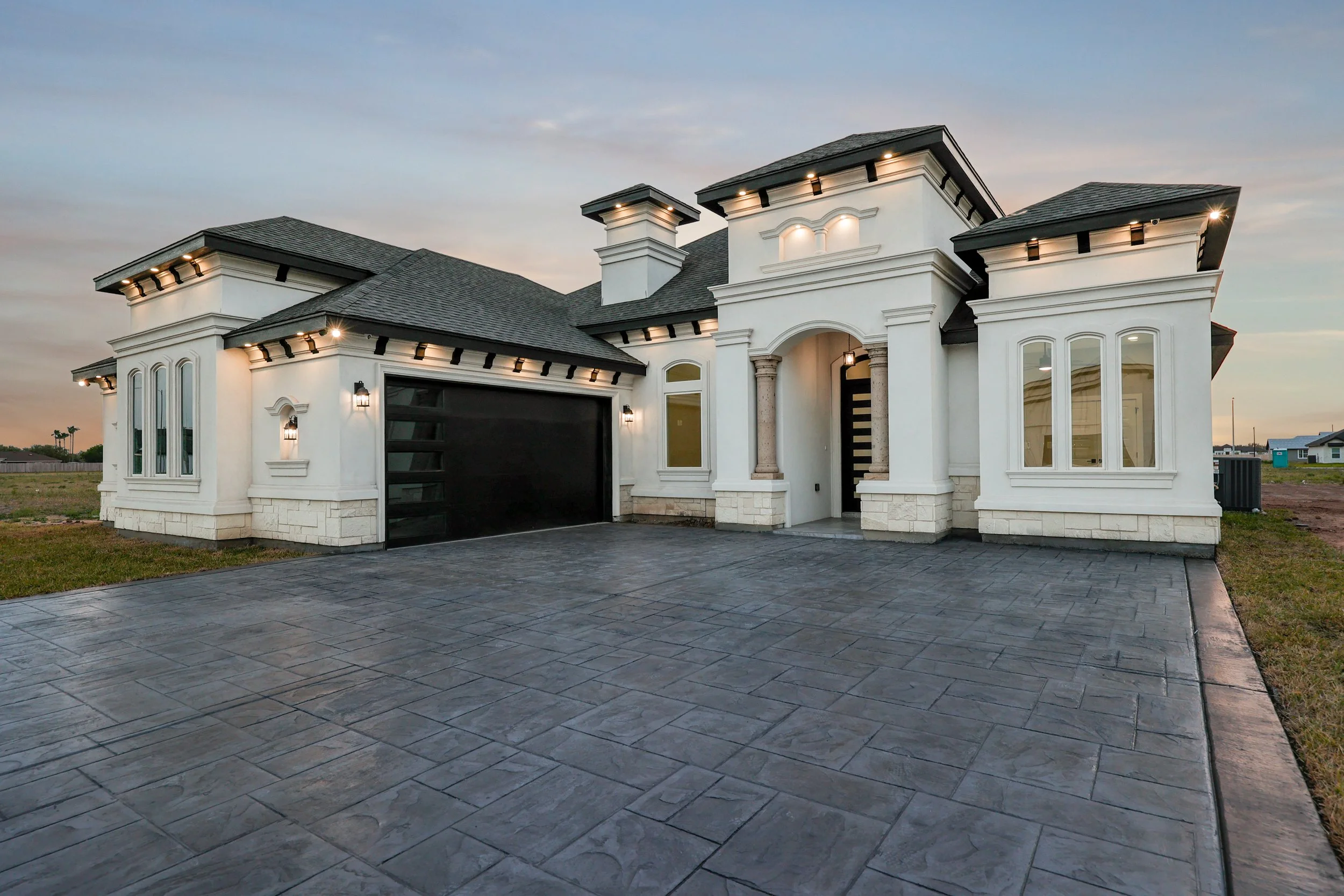 Modern white stucco house with a large driveway, dark roofing, and outdoor lighting at twilight.
