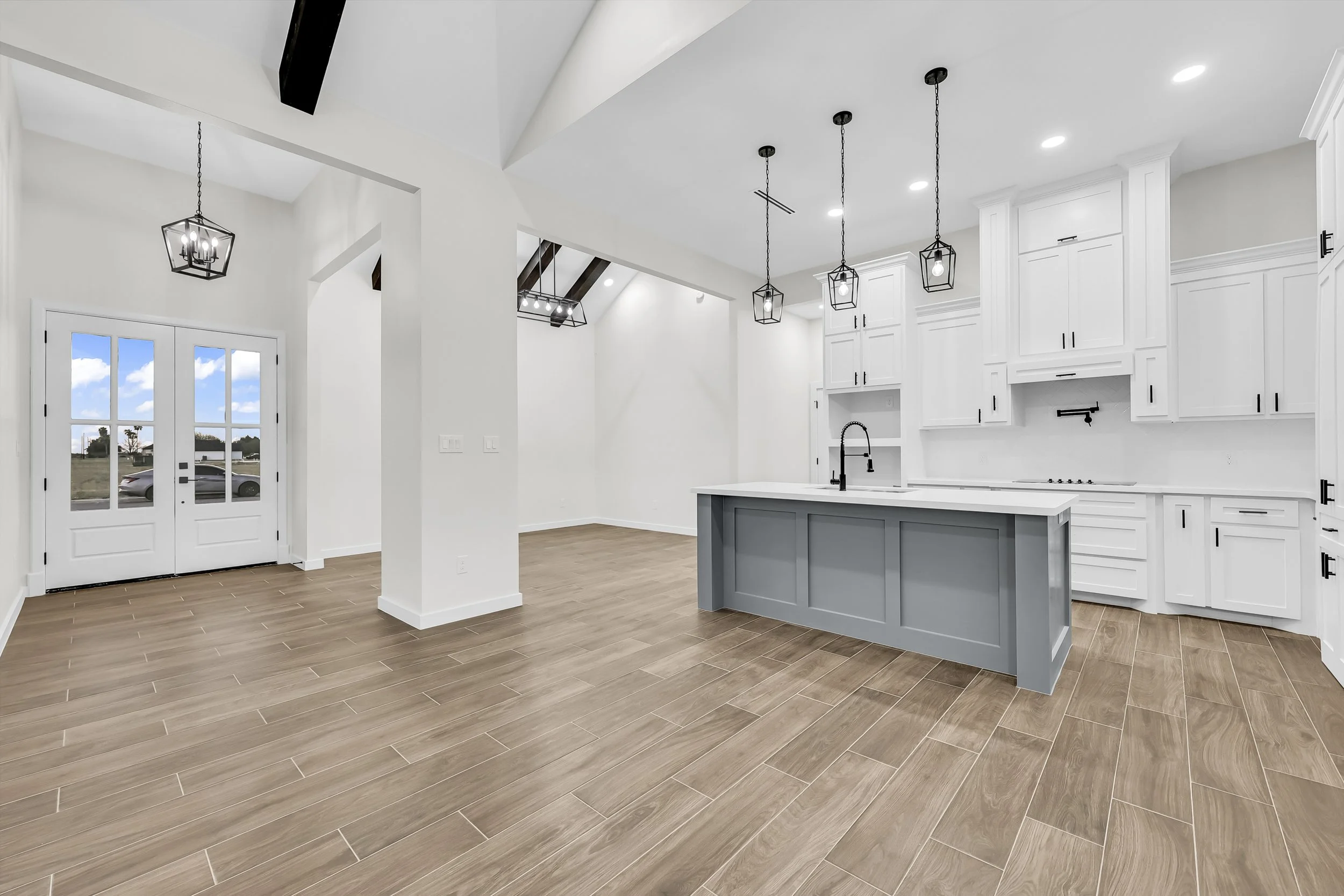 Modern kitchen interior with white cabinetry, gray kitchen island, and wood-look tile flooring. Features pendant lights and a double front door with glass panels.