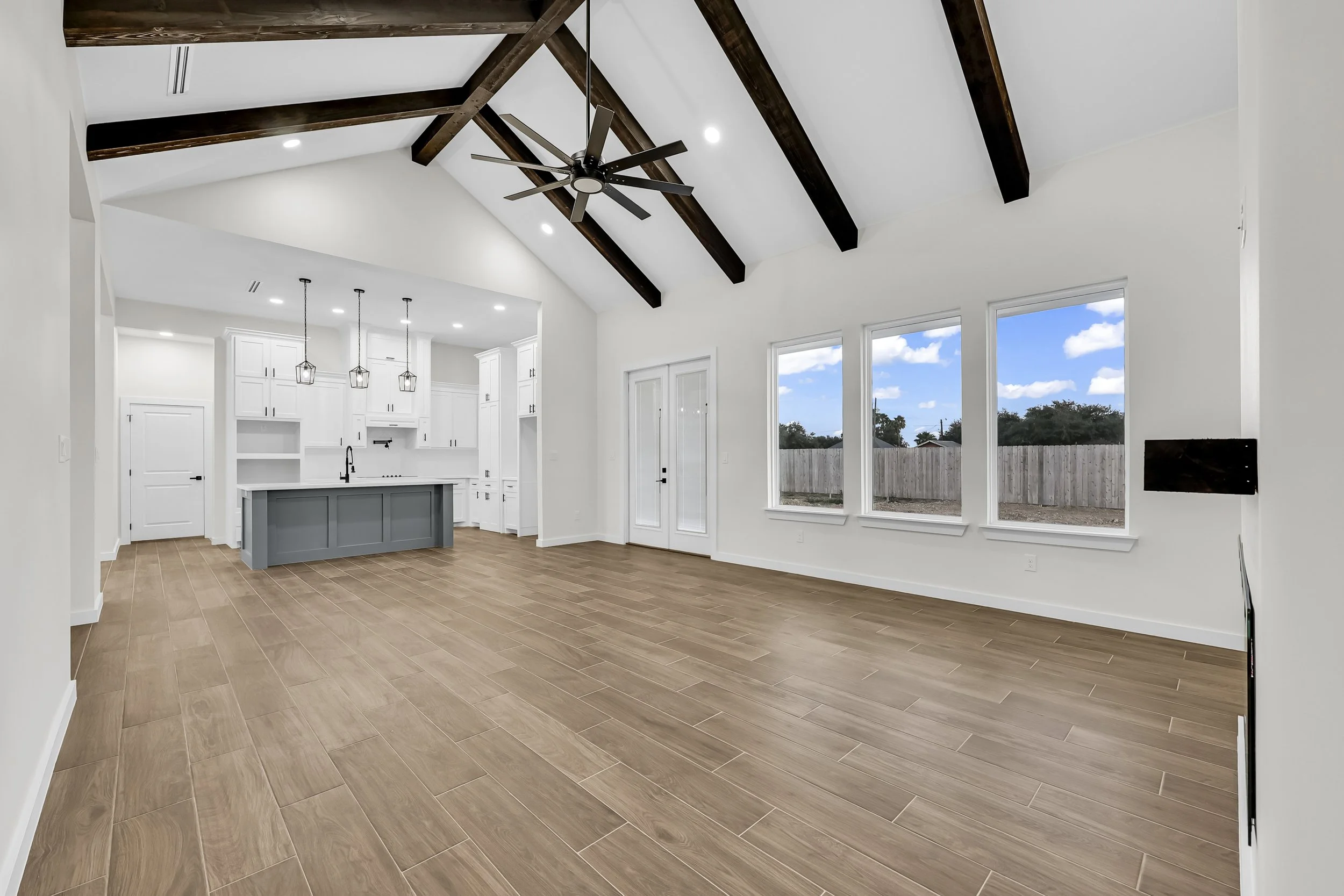 Spacious modern interior with high vaulted ceiling, exposed wooden beams, and a ceiling fan. The room features a light wooden floor, large windows offering natural light, and a white open kitchen with a center island and pendant lights. Double glass 