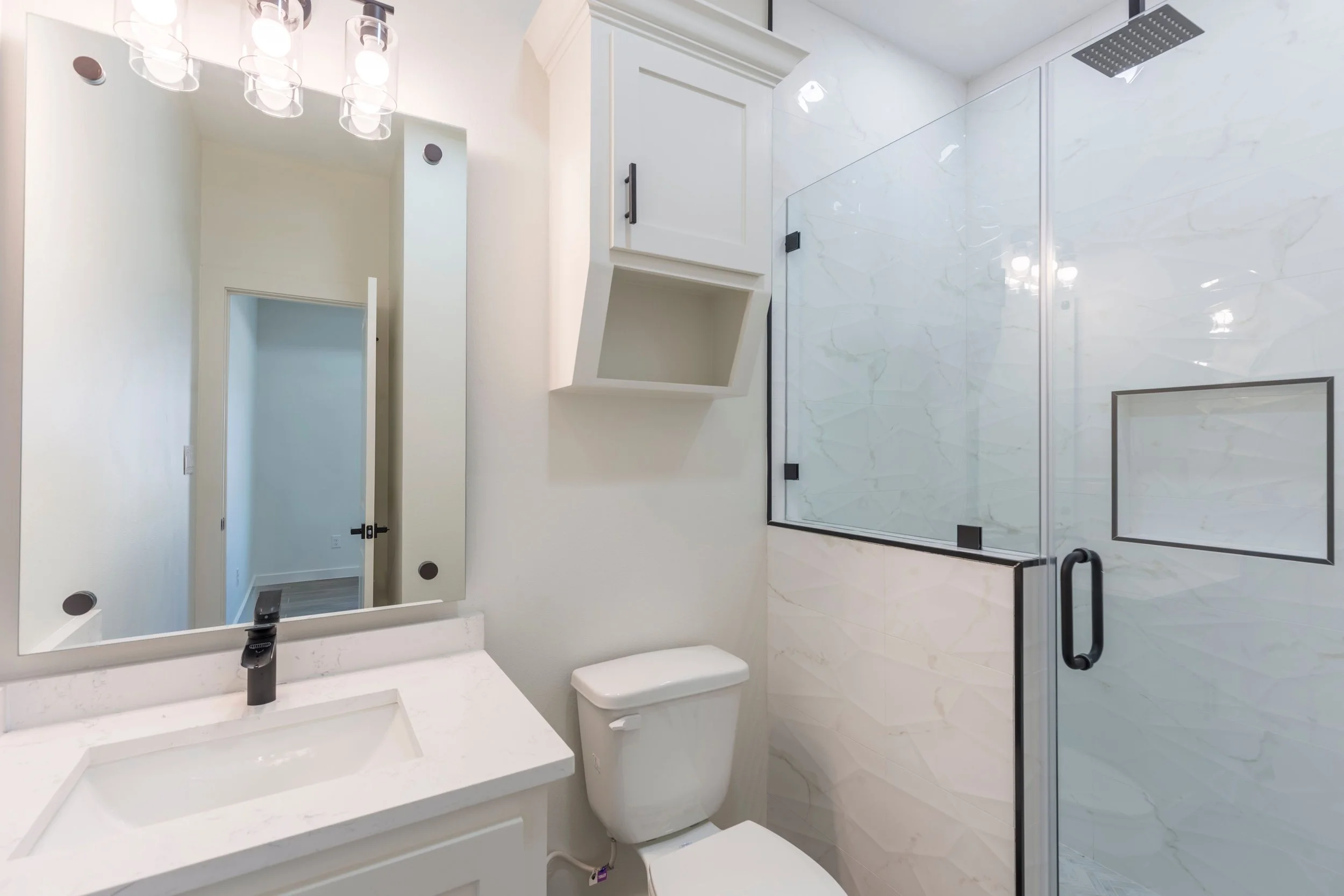 Modern bathroom with white vanity, sink, toilet, and glass shower enclosure. Large mirror with lights overhead, light-colored walls, and geometric wall tiles.
