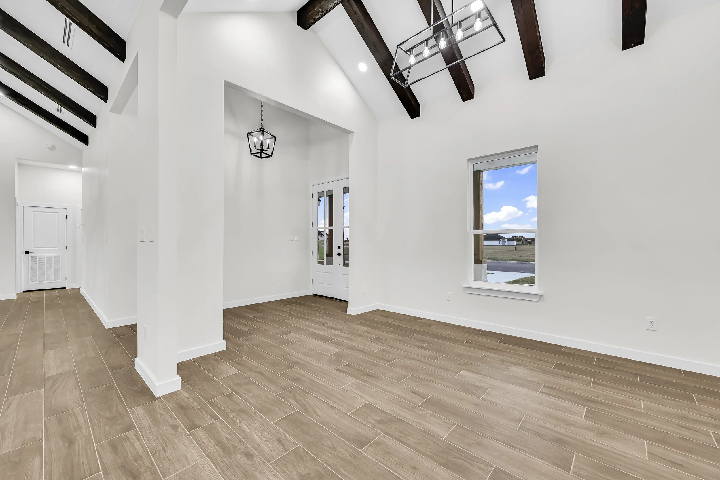 Modern empty room with high ceilings, wood-look tile flooring, exposed wooden beams, white walls, large window, and two geometric pendant lights.