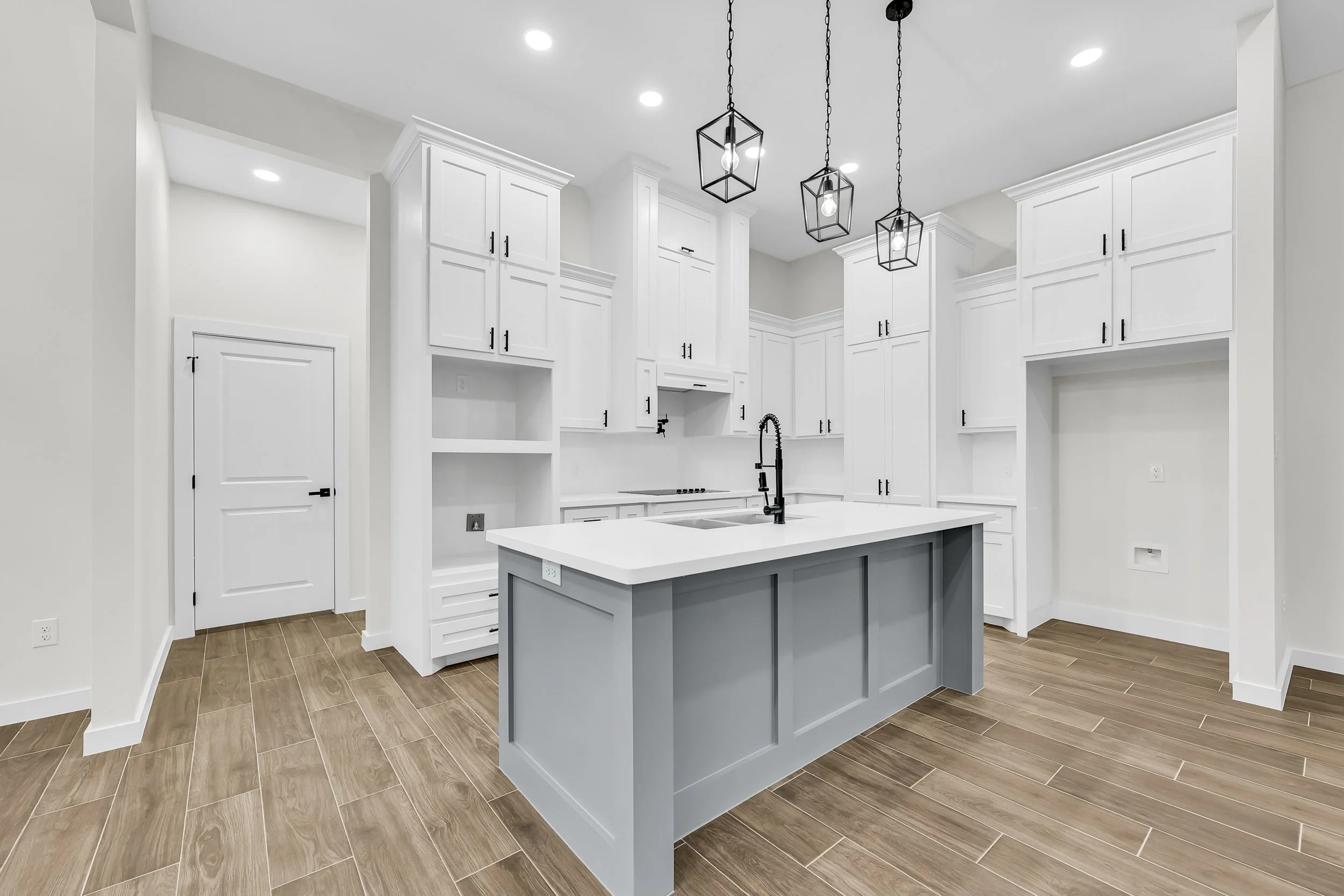 Modern kitchen interior with white cabinets, large island with gray base, pendant lighting, and wood-style tile flooring.