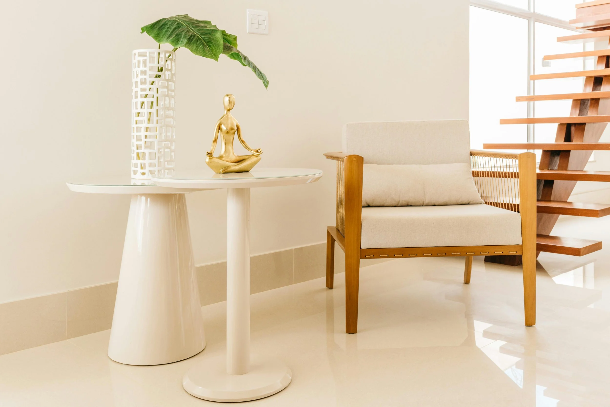 Minimalist living space with a white round side table holding a tall white decorative vase with a large green leaf, and a small gold figurine of a person meditating. There is a beige armchair with wooden armrests and a wooden stairway leading to an upper level.