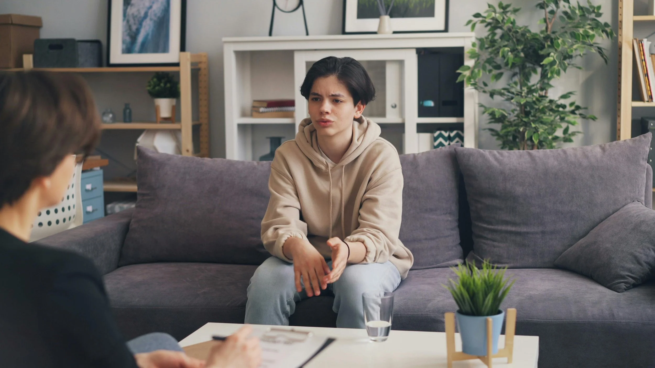 Young woman with short dark hair wearing a beige hoodie and light-colored pants sitting on a gray couch, having a conversation in a living room. The room has a white fireplace mantle, framed pictures, green plants, and wooden shelves with decor.