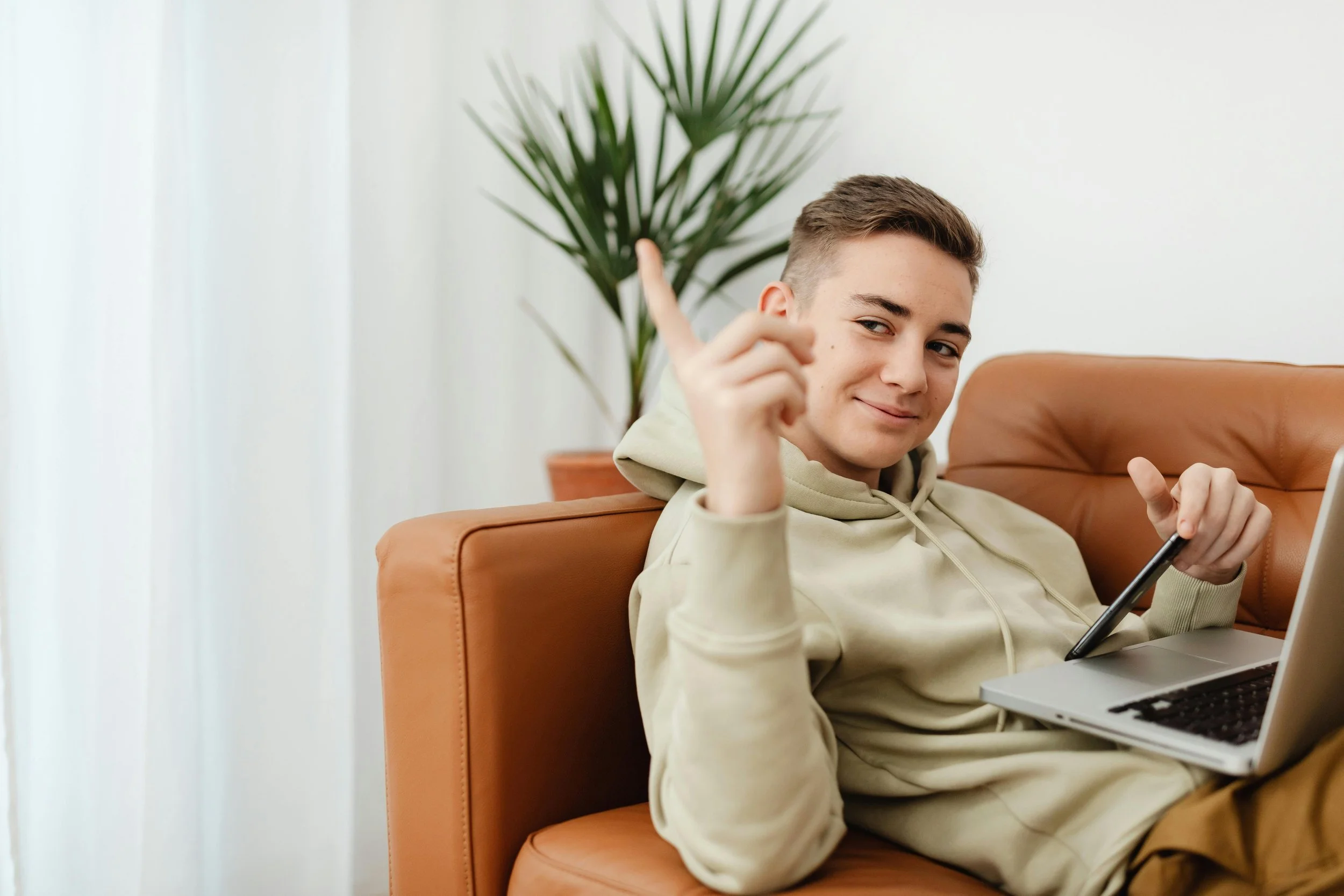 A young man with short brown hair sitting on an orange couch, holding a smartphone in one hand, pointing upward with the other, smiling, with a large green plant behind him in a bright room.