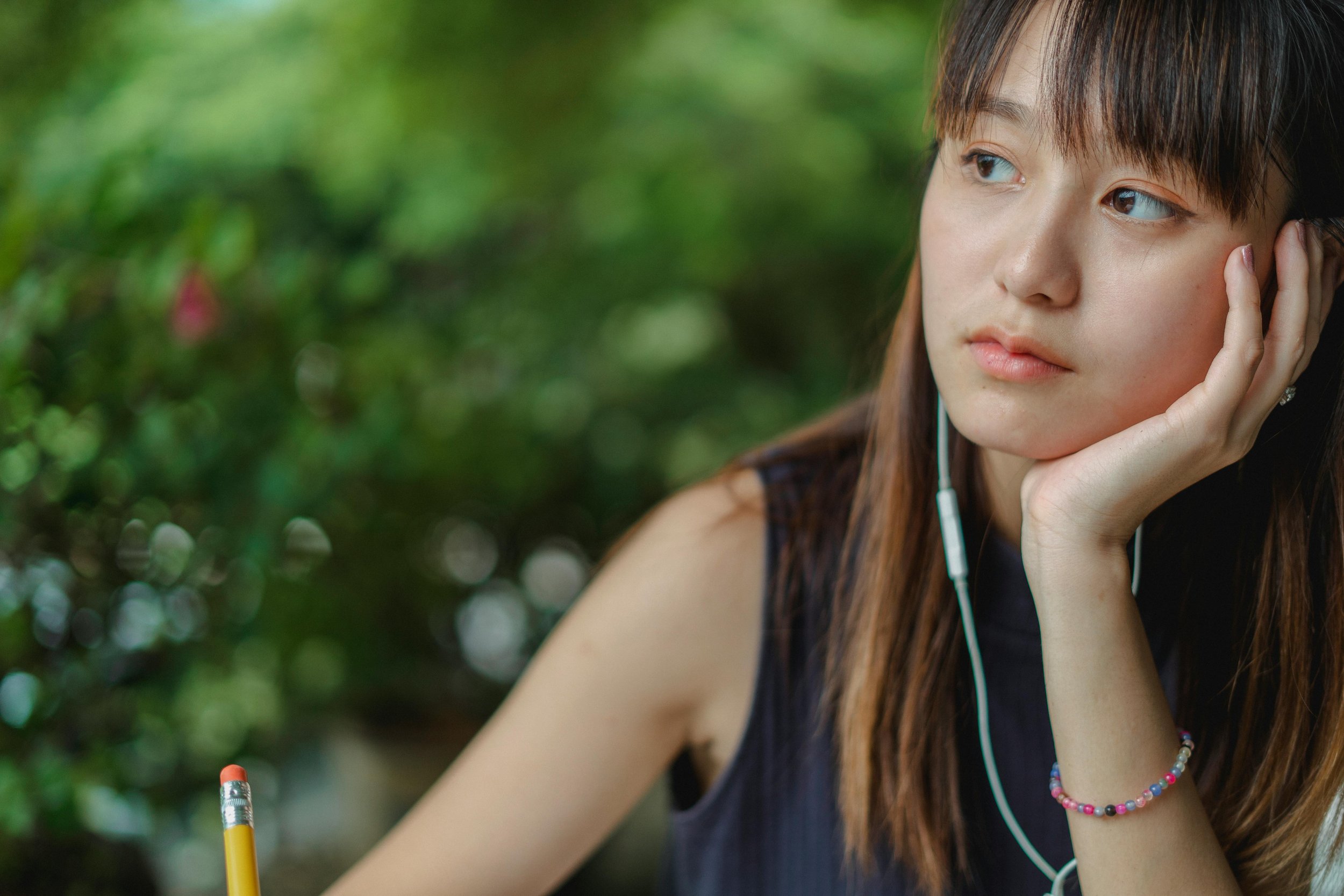 A young woman with long brown hair, wearing a beaded bracelet and necklace, resting her face in her hand with an earbud in her ear, outdoors with a green blurred background.