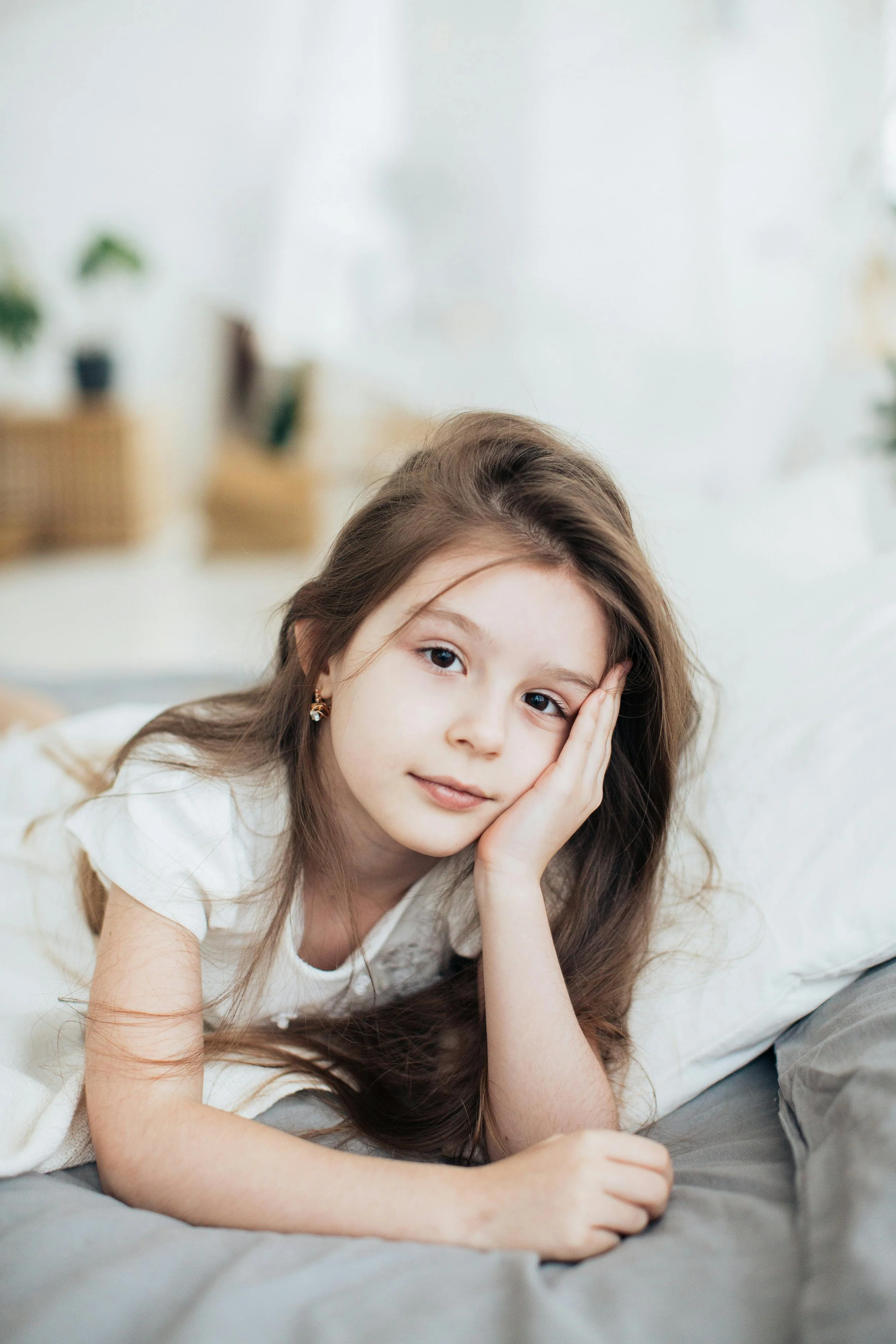 A young girl with long brown hair lying on a bed, resting her head on her hand, looking ahead thoughtfully in a well-lit room with minimal decor and houseplants in the background.