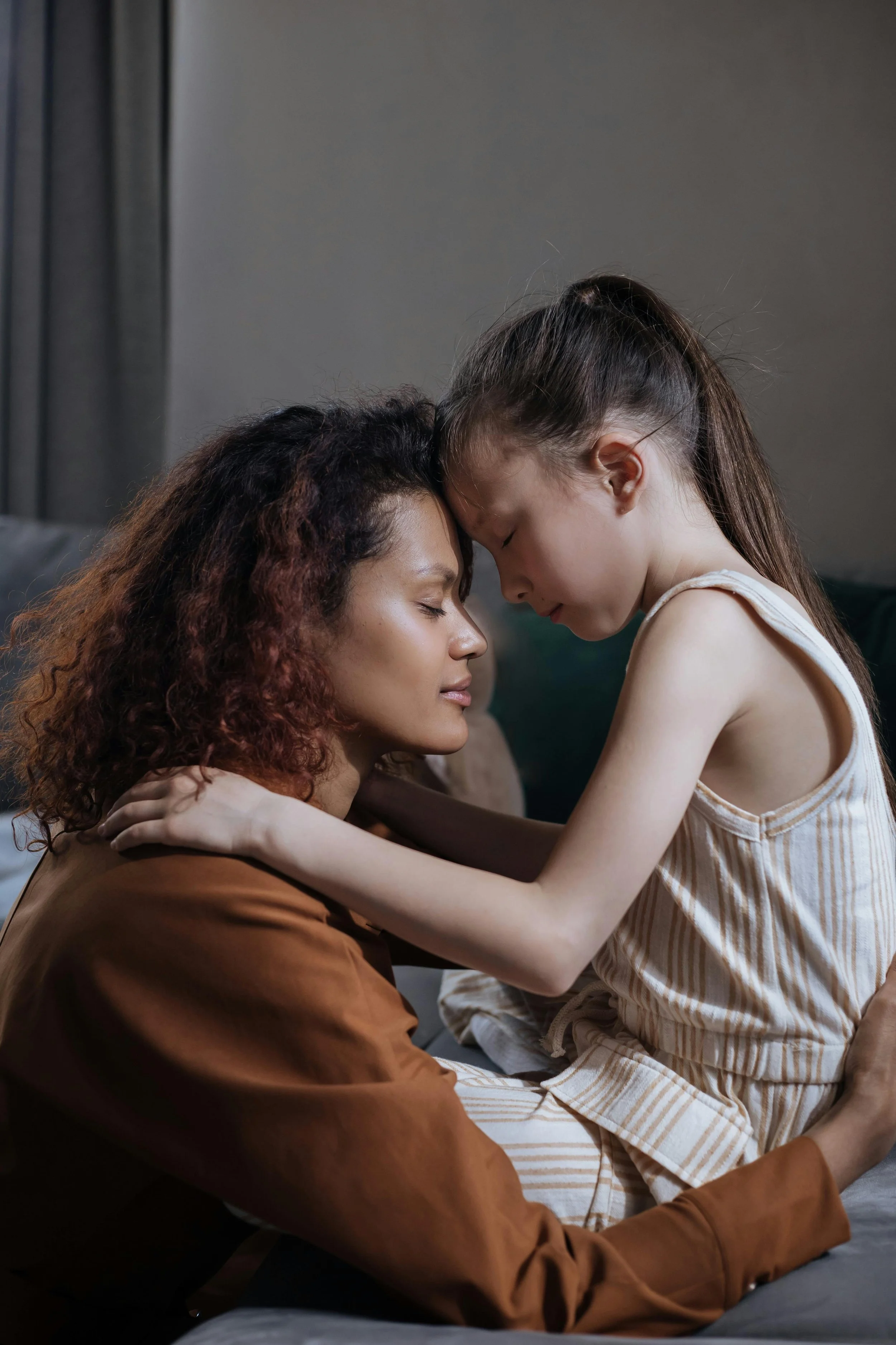 A woman and a young girl, likely mother and daughter, sharing a tender moment with foreheads touching, eyes closed, and arms wrapped around each other, in a cozy indoor setting.