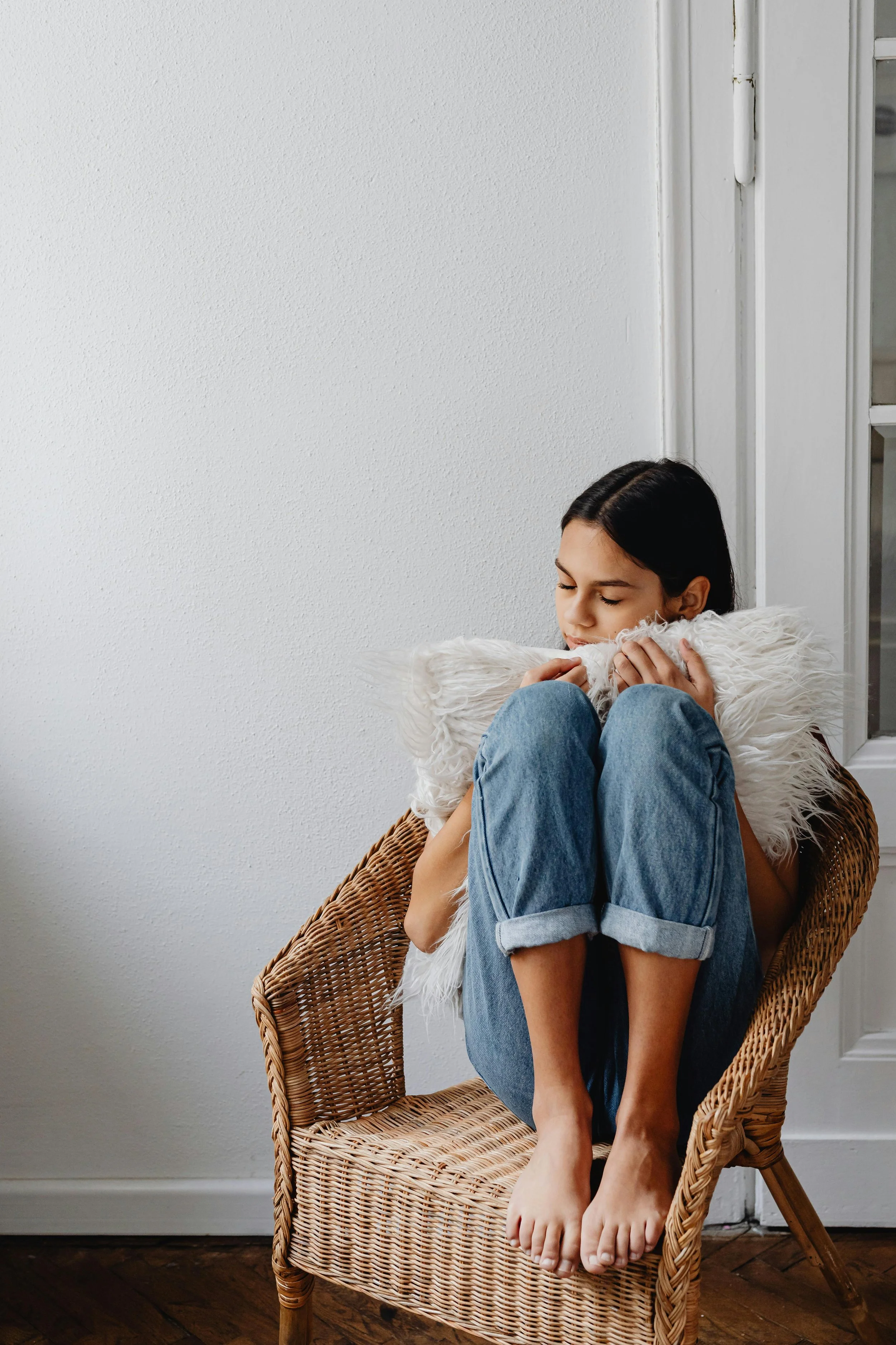 A young woman sitting in a wicker chair, holding a fluffy white pillow, with her eyes closed and head tilted downward. She wears a yellow top and ripped blue jeans, sitting barefoot, indoors near a window with white trim.
