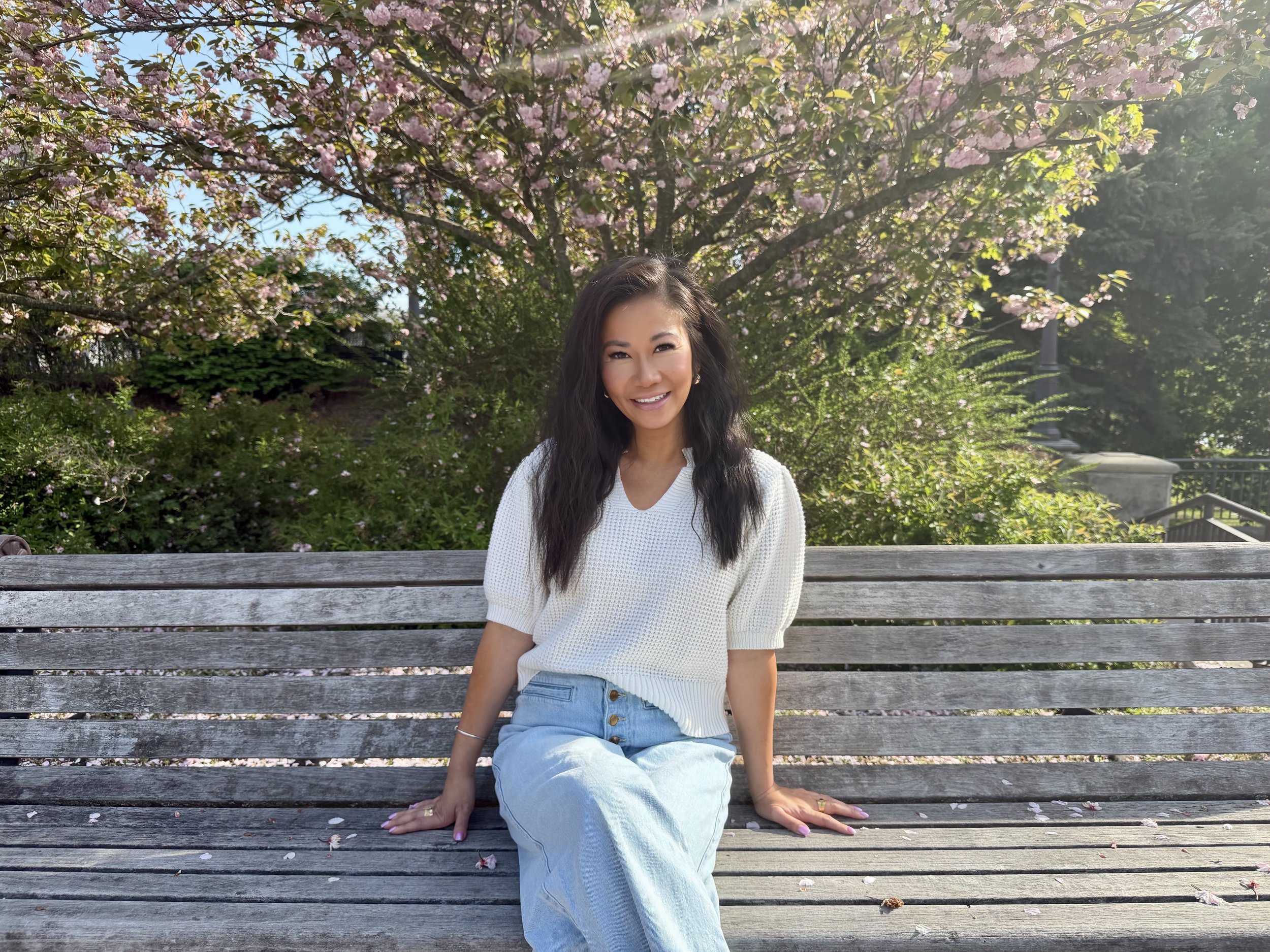 A woman with long dark hair smiling while sitting on a wooden park bench under a flowering tree with pink blossoms, wearing a white sweater and light blue jeans, with sunlight filtering through the leaves.