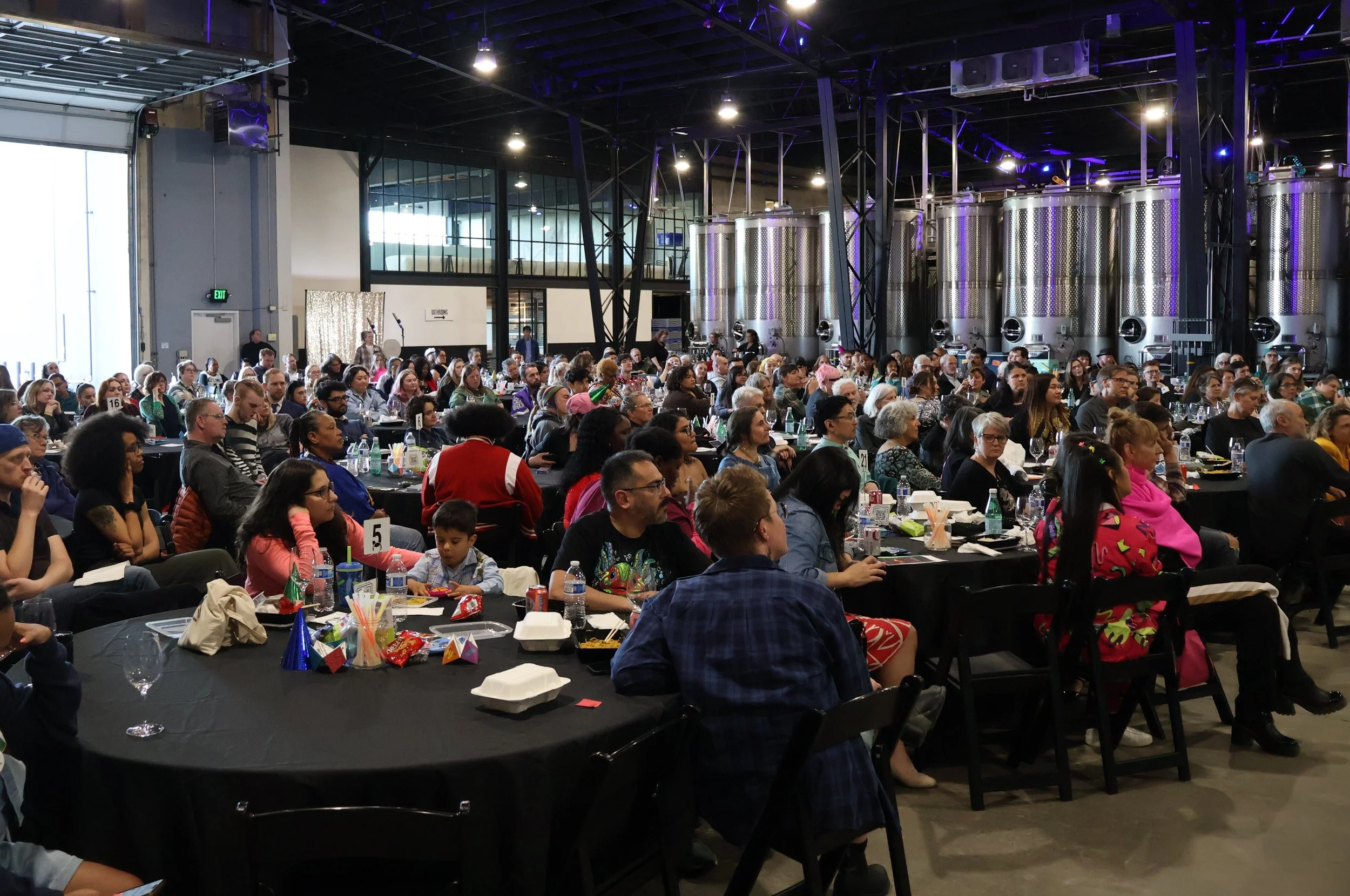 Photo of crowded room filled with hundreds of guests at an event in a brewery, listening raptly to the speaker